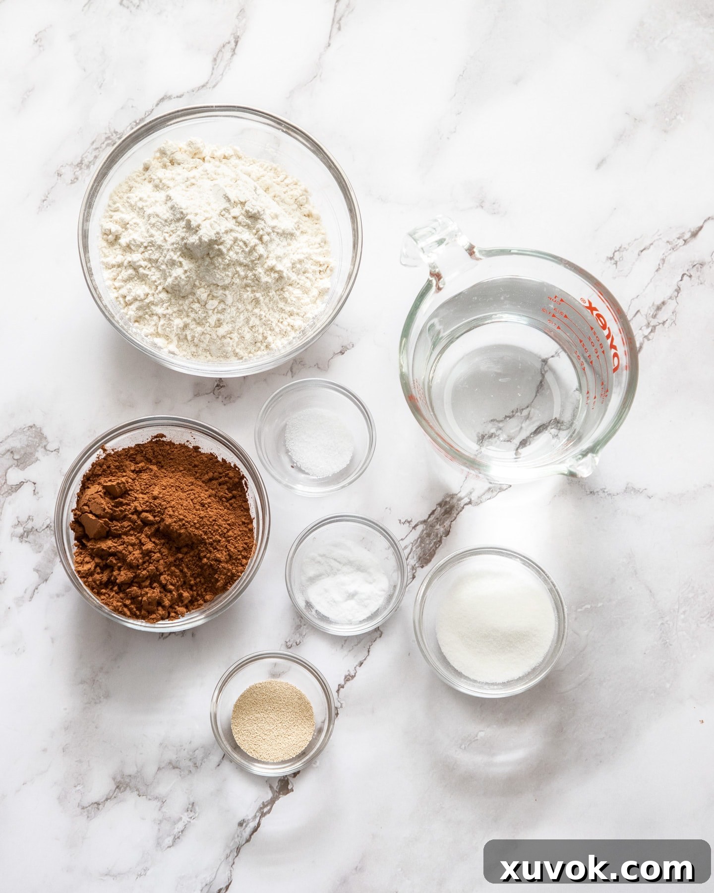 Ingredients for chocolate crumpets laid out on a marble surface, including flour, cocoa powder, sugar, yeast, baking soda, salt, and water. All ingredients are clearly visible and organized.