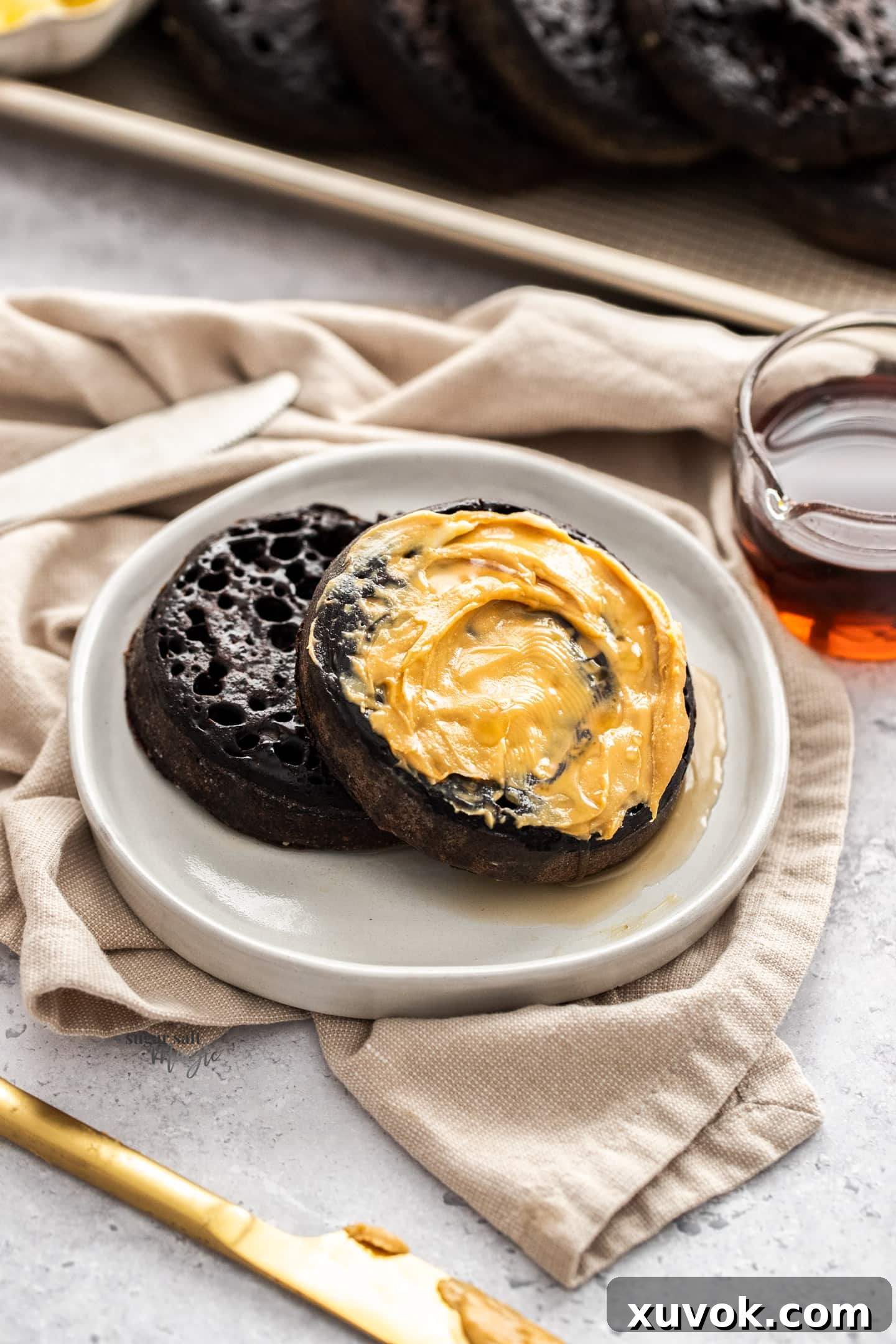 Two chocolate crumpets on a small plate, generously smothered in peanut butter and drizzled with maple syrup. The crumpets are warm and inviting, showcasing their porous texture.