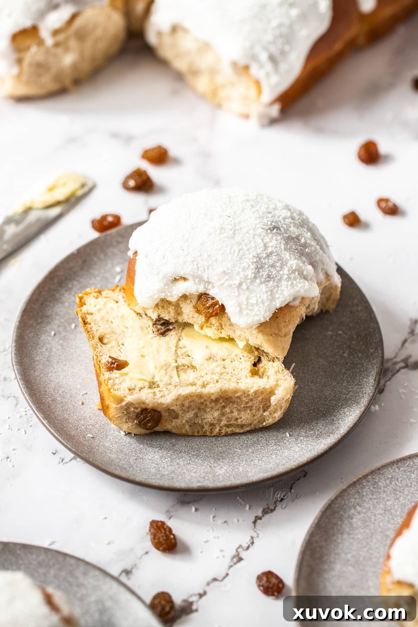 An iced bun on a grey dessert plate with sultanas scattered around.