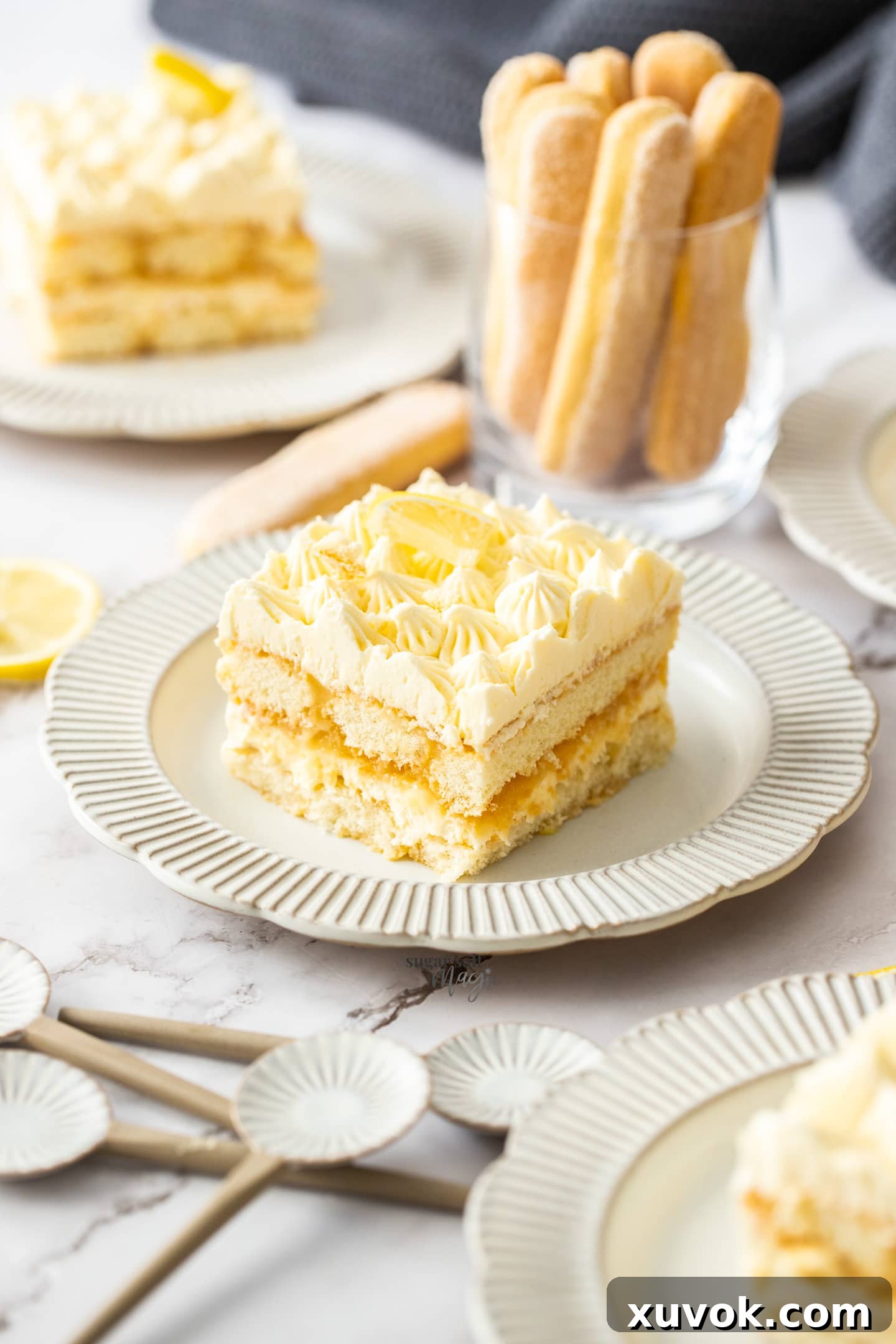 A square slice of lemon tiramisu on a small plate with a cup of lady fingers the background.