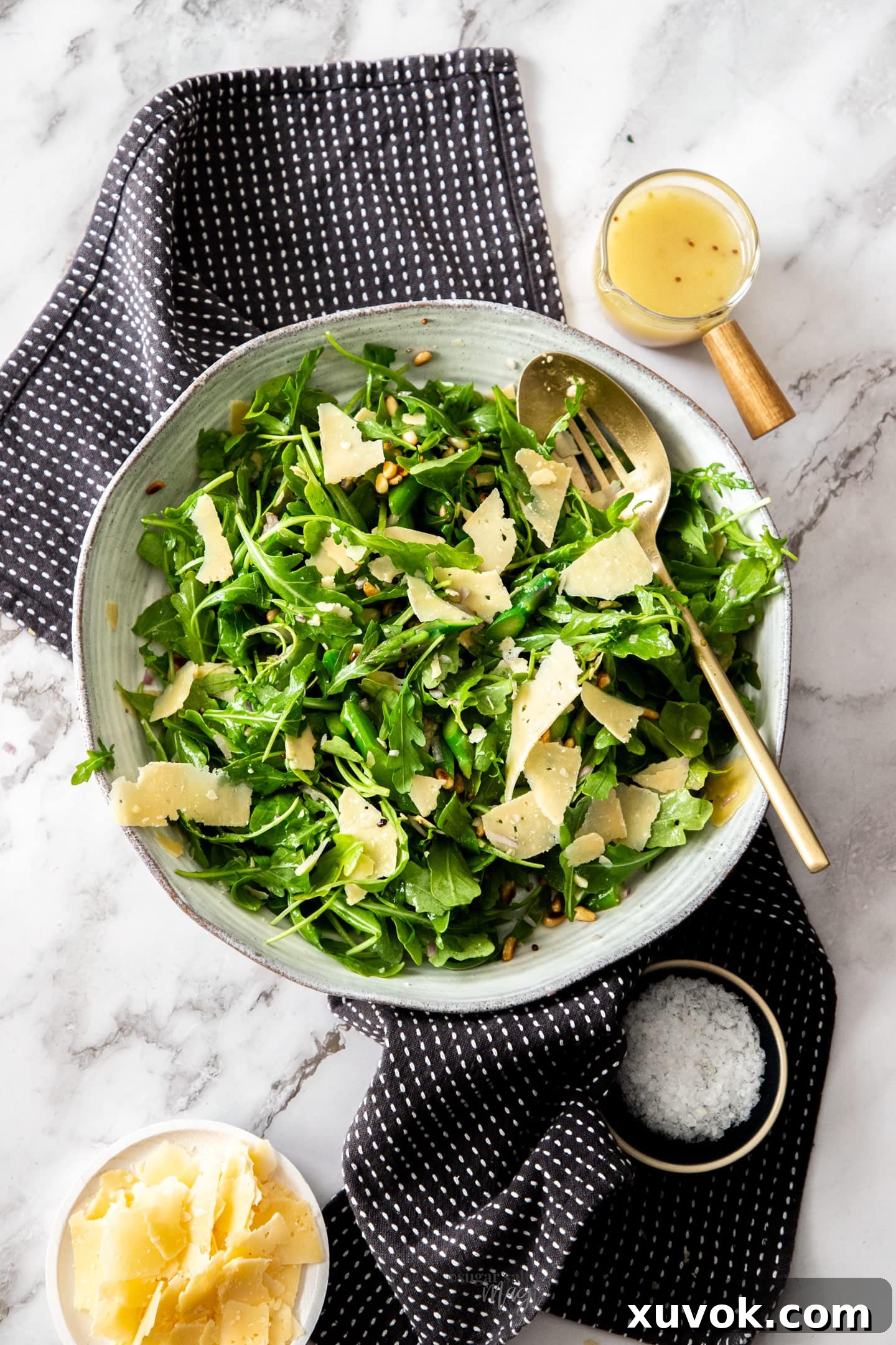 A large bowl filled with vibrant green asparagus, rocket, and parmesan salad with a small pot of lemon mustard dressing on the side.