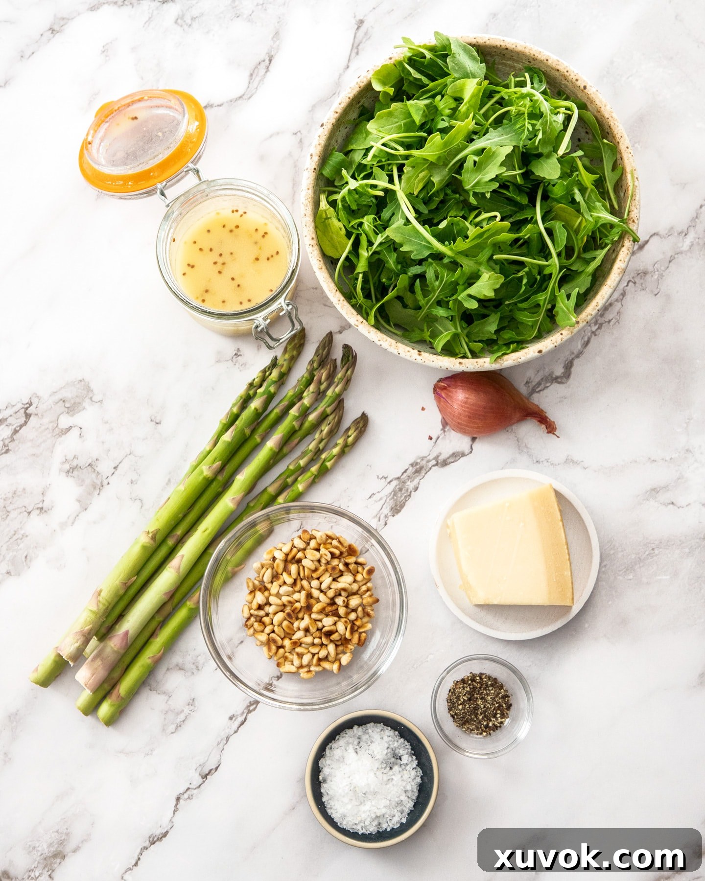 Ingredients for rocket and parmesan salad laid out on a marble benchtop including asparagus, rocket, parmesan block, pine nuts, shallot, and dressing components.