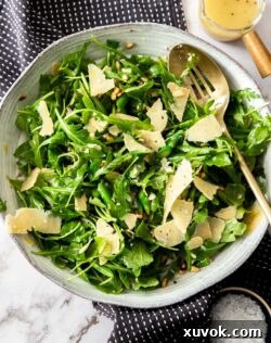 A large bowl filled with vibrant green asparagus, rocket, and parmesan salad with a small pot of dressing on the side.