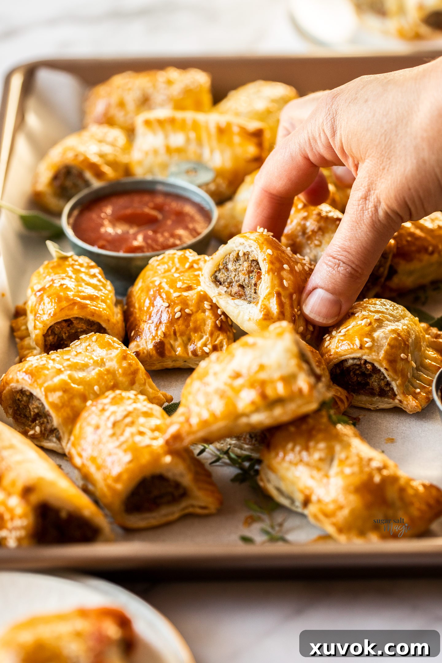 A hand reaching for a perfectly golden-brown turkey sausage roll from a large batch on a baking tray.