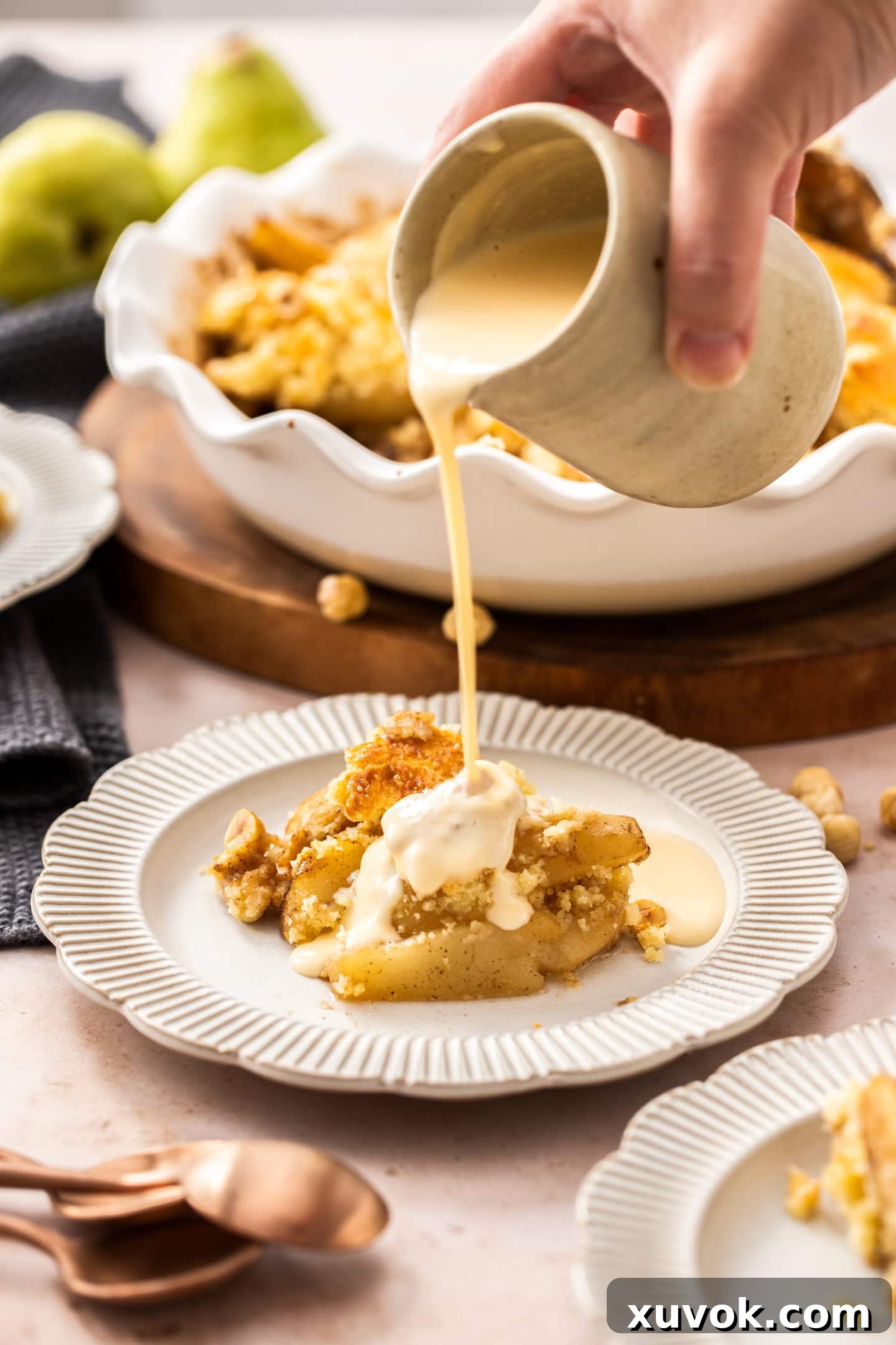Custard being poured onto a serving of pear cobbler on a dessert plate, highlighting the warm fruit and topping.