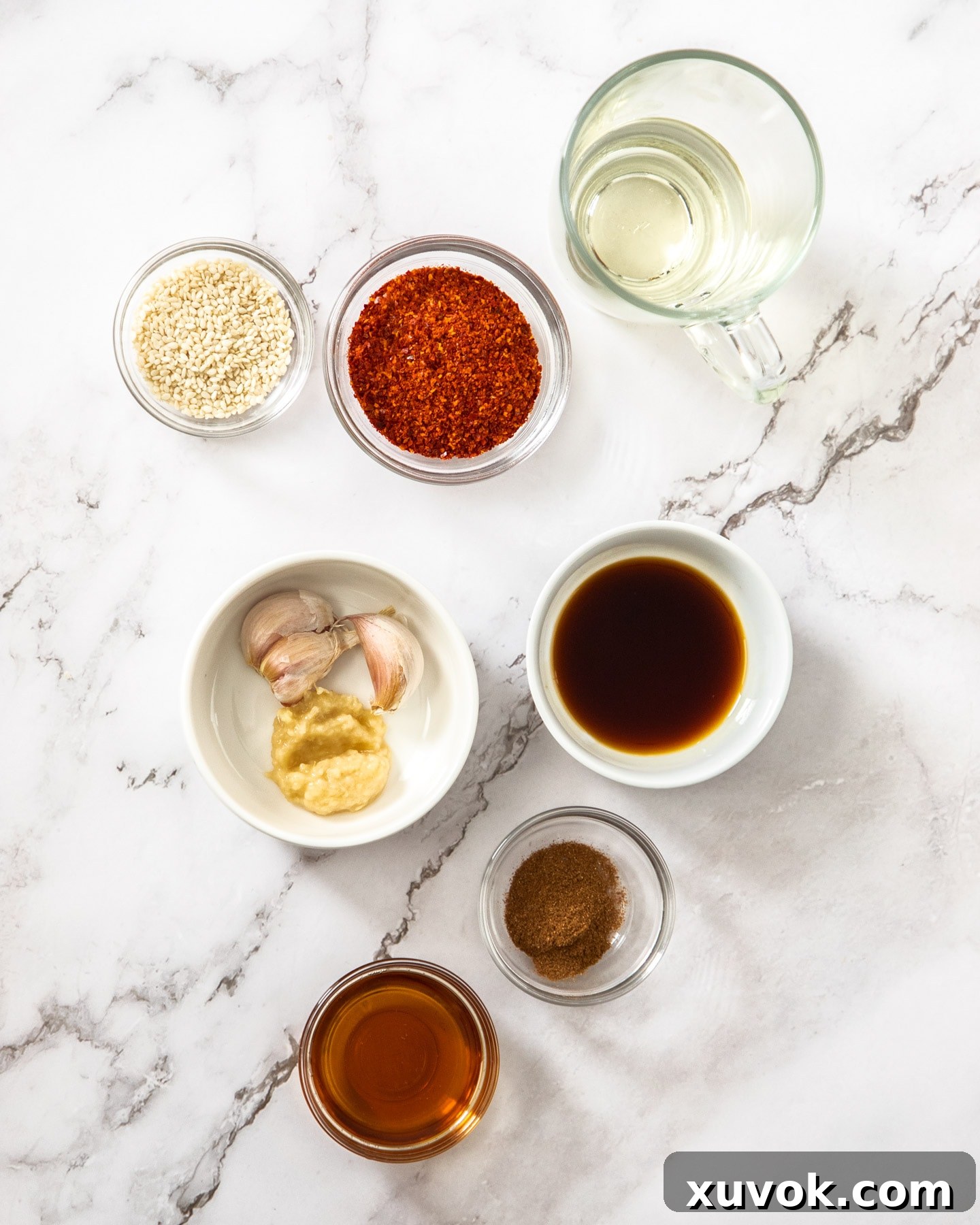 Ingredients for sesame chilli oil arranged on a marble surface, including sesame oil, garlic, Korean chilli flakes, sesame seeds, ginger, five spice powder, and soy sauce.