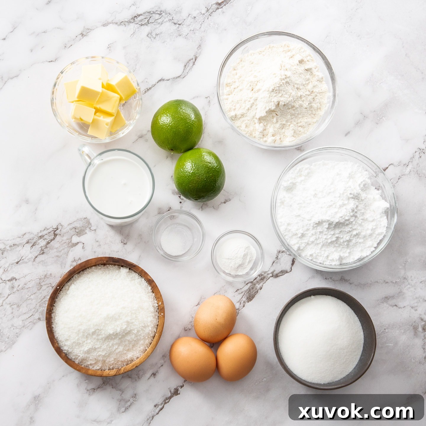 A flat lay of fresh ingredients for lime and coconut cake, including limes, coconut cream, flour, and butter, arranged neatly on a marble surface.