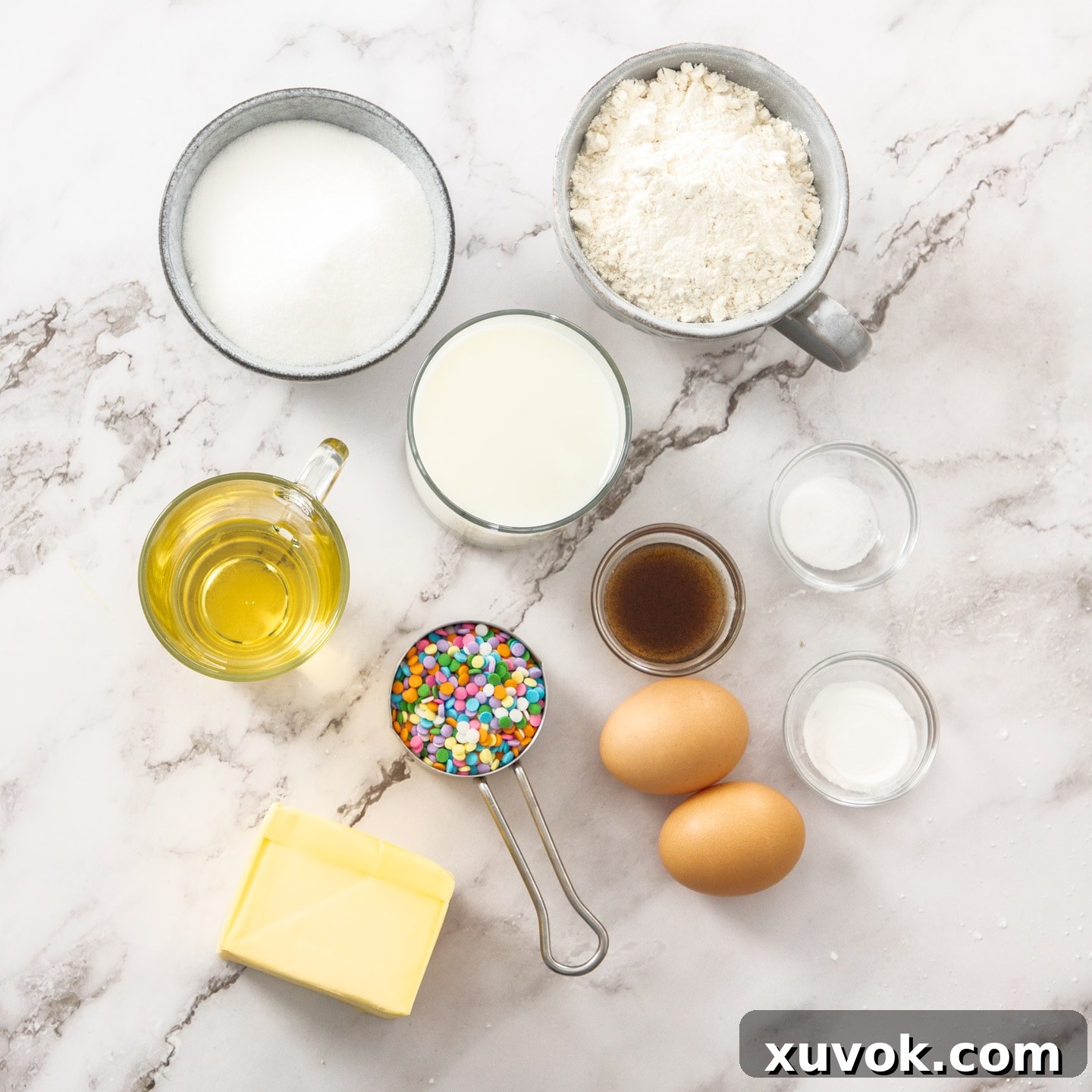 Ingredients for confetti cupcakes, including flour, sugar, butter, eggs, and colorful sprinkles, laid out on a marble surface.