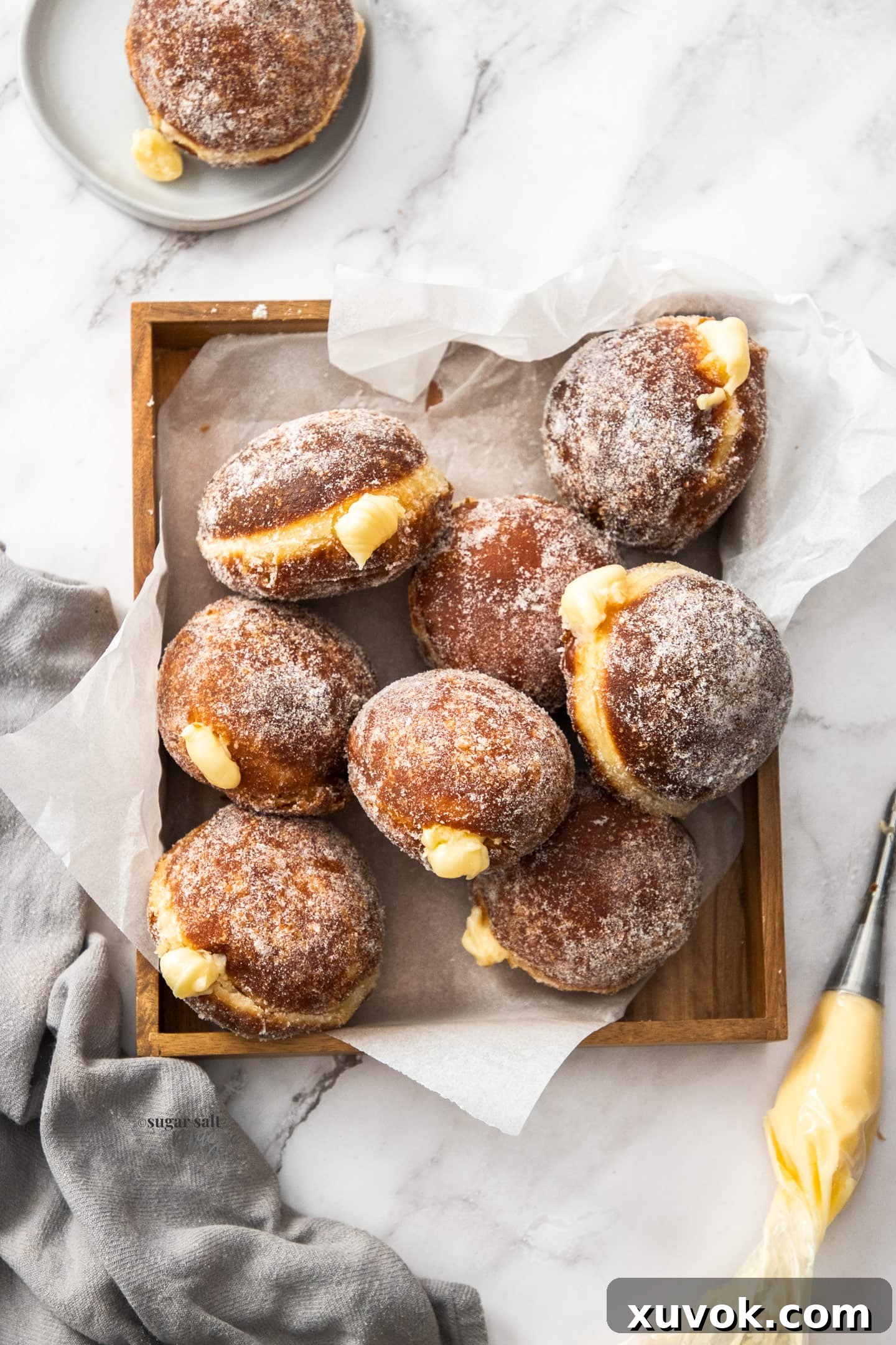 Eight perfectly sugar-coated Bavarian cream donuts arranged in a wooden tray, with a piping bag filled with cream ready to finish the last one.