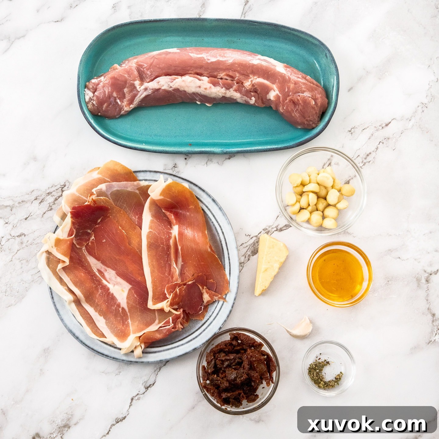 A flat lay photograph of the key ingredients required for making stuffed pork tenderloin, including pork, prosciutto, sundried tomatoes, macadamias, garlic, parmesan, and herbs.