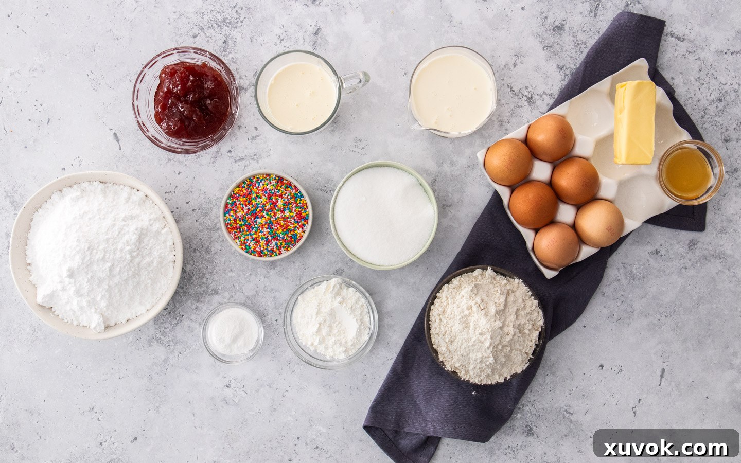 Ingredients for fairy bread lamingtons laid out on a grey stone bench top, including butter, milk, vanilla, eggs, flour, baking powder, salt, sugar, cream, jam, and sprinkles.