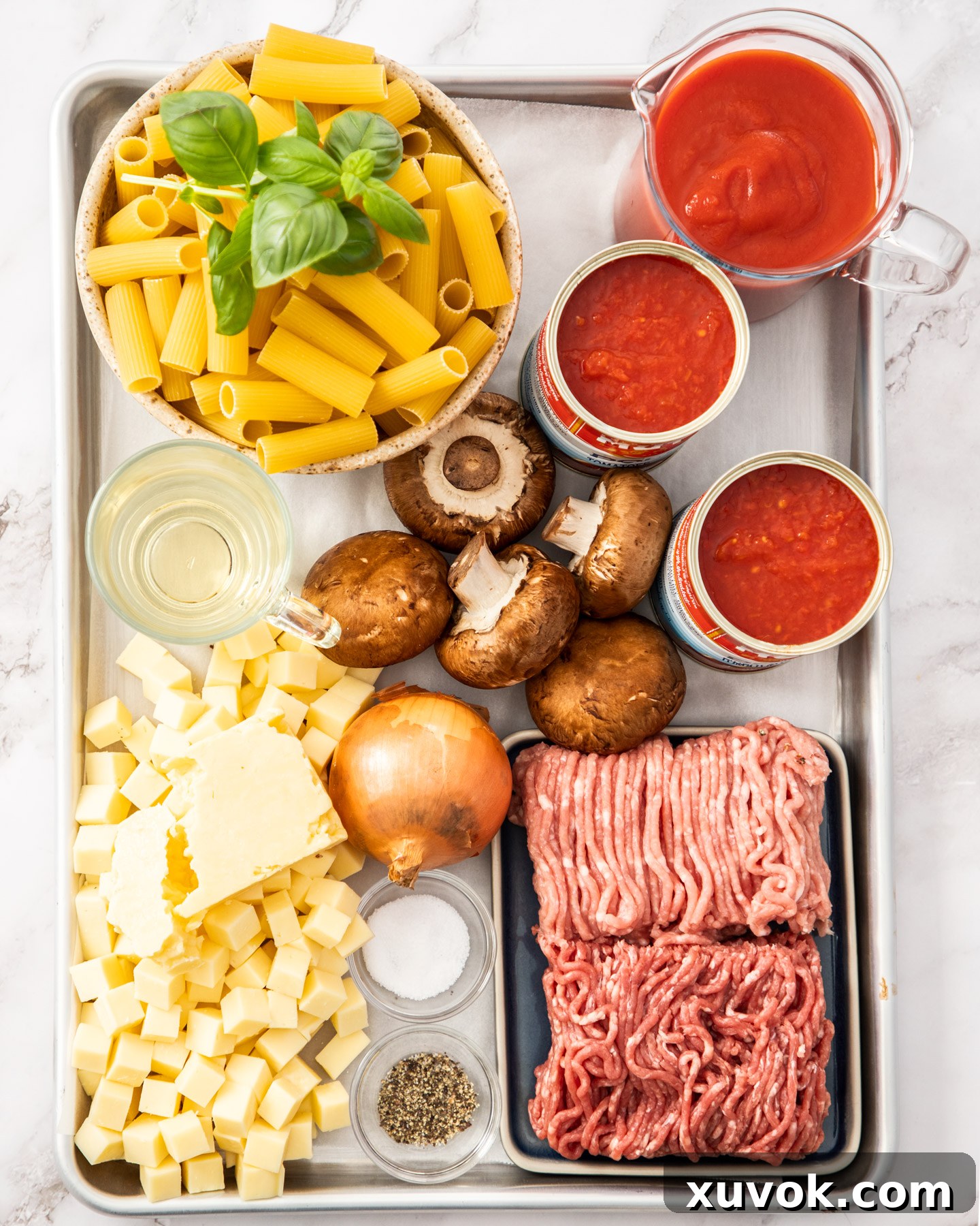 A collection of fresh ingredients for rigatoni al forno laid out on a baking tray, including onions, mushrooms, ground meats, canned tomatoes, pasta, and cheeses.