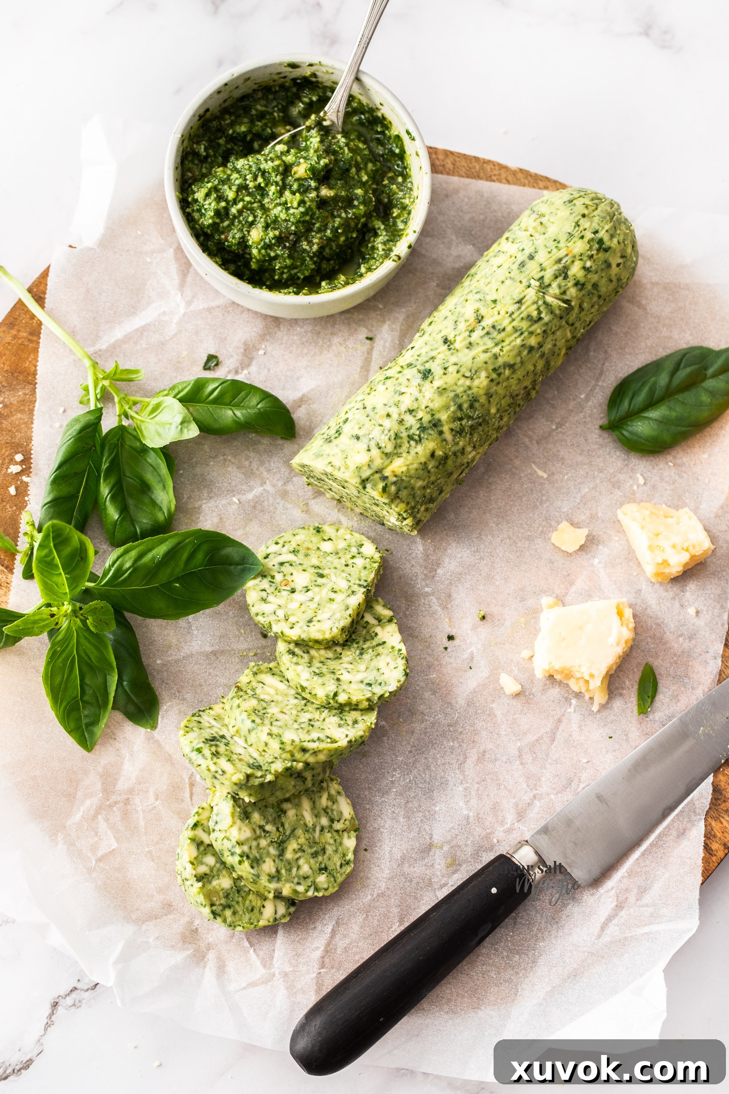 A log of beautifully marbled green basil pesto butter on a rustic chopping board, with several disks already cut and ready for use. Fresh basil leaves and grated Parmesan are scattered around.