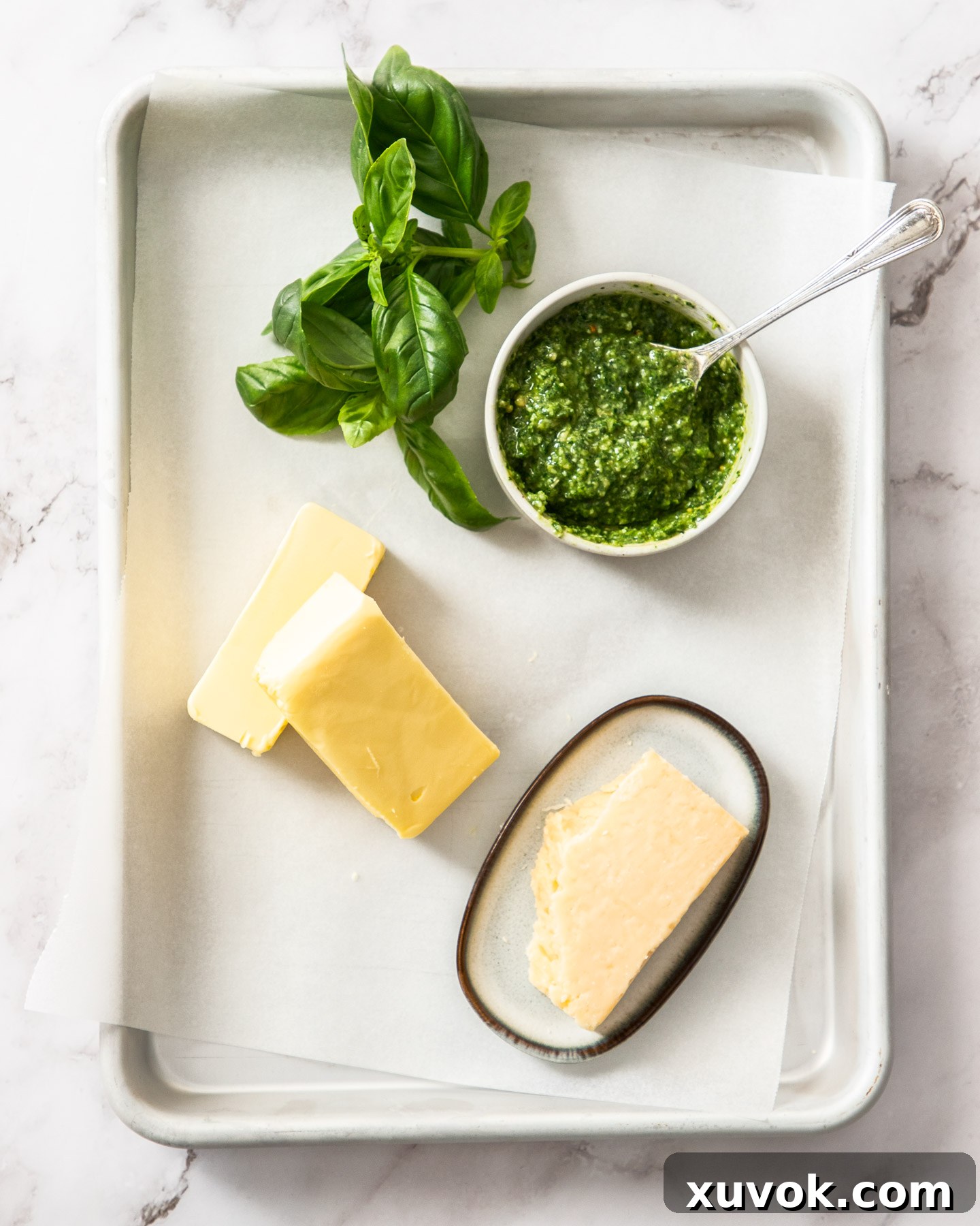 All the fresh ingredients for basil pesto butter laid out on a baking tray, including unsalted butter, vibrant basil pesto, block Parmesan cheese, and fresh garlic cloves.