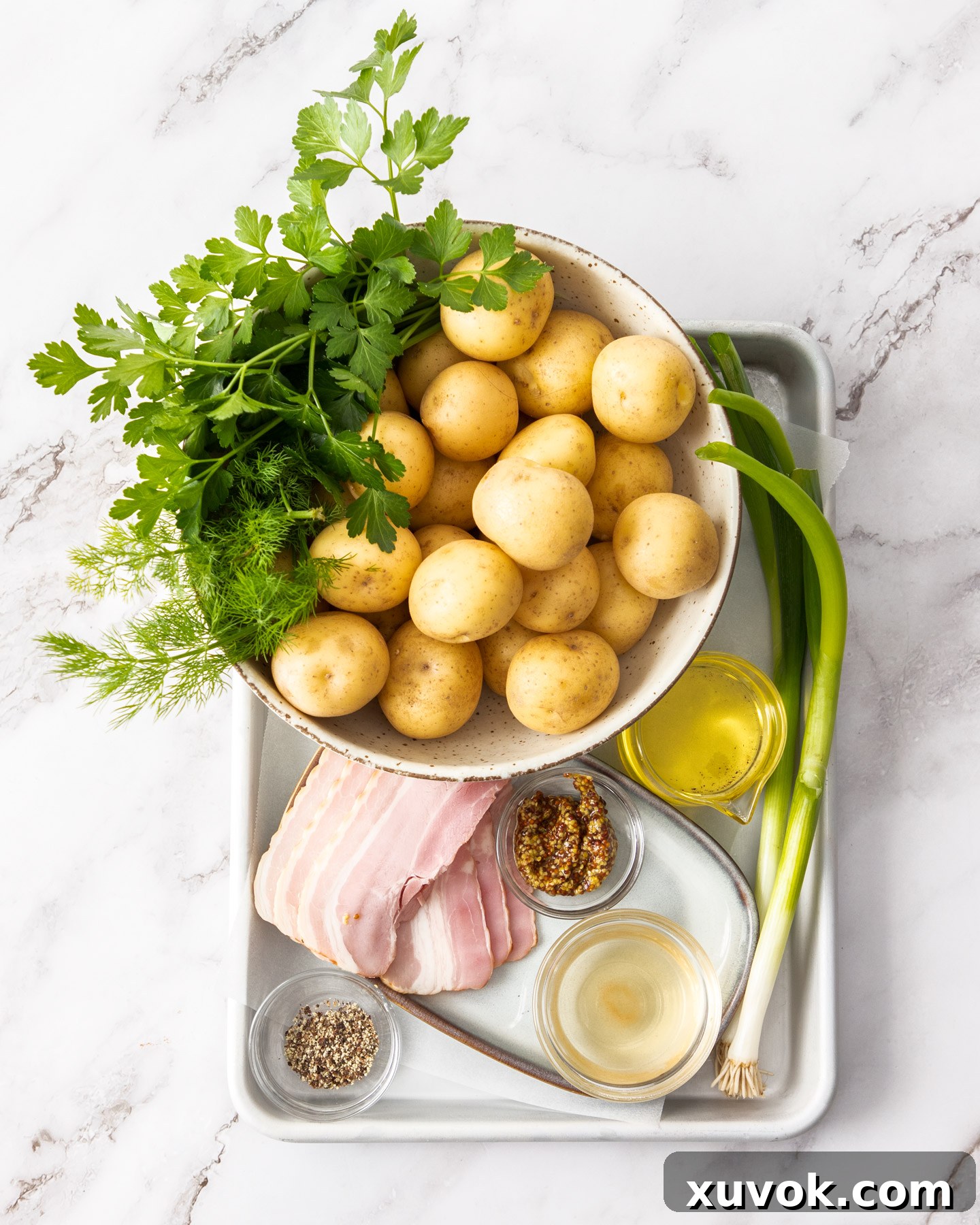 A collection of fresh ingredients laid out, including baby potatoes, bacon, spring onions, fresh herbs, olive oil, and mustard, ready for making mayo-free potato salad.