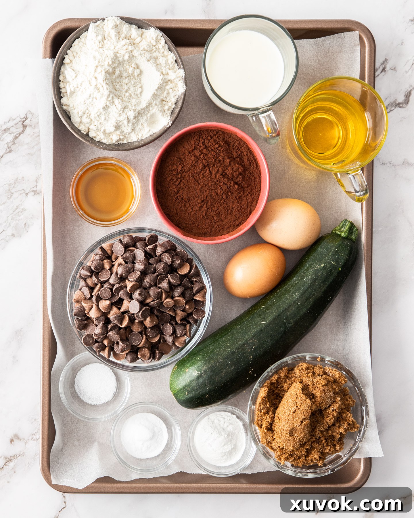 All the necessary ingredients for baking a chocolate zucchini cake laid out on a baking tray, including flour, cocoa, sugar, zucchini, and chocolate chips.