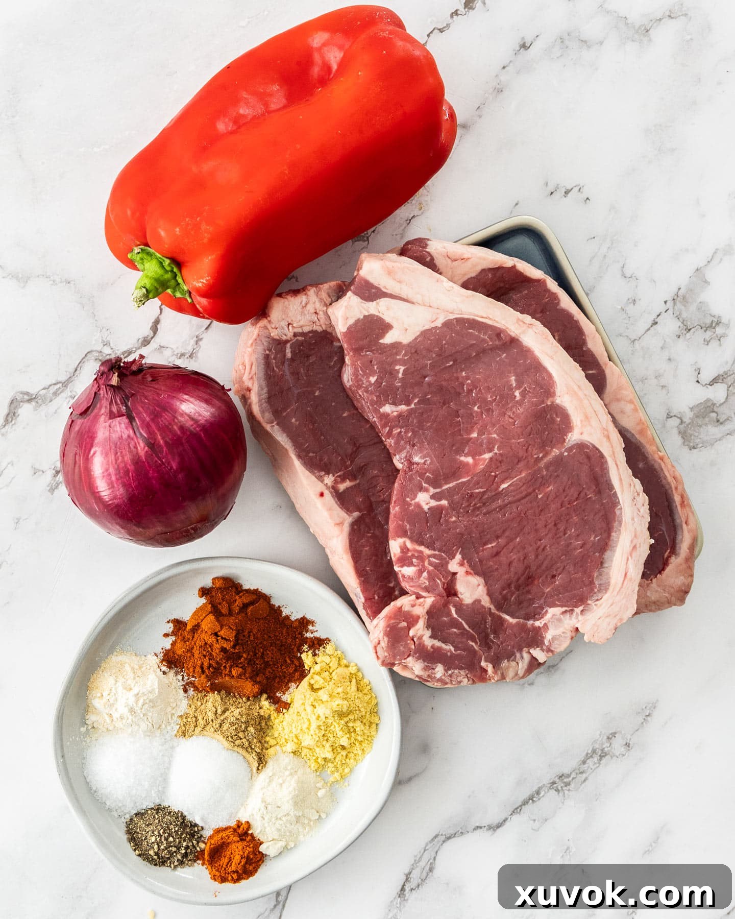 Ingredients for beef kabobs laid out on a marble surface, including meat, vegetables, and spices.