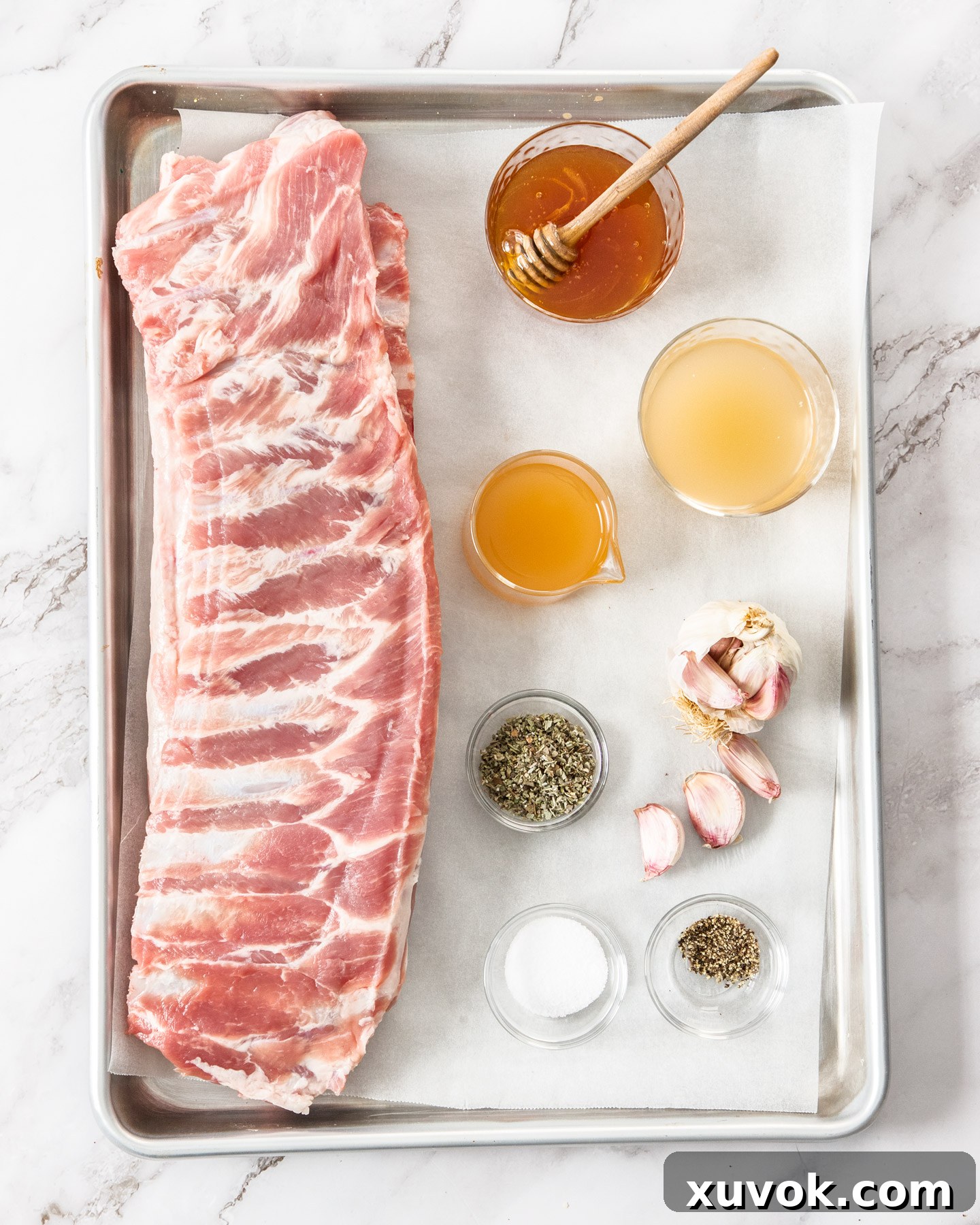 All the essential ingredients for making honey garlic pork ribs, neatly arranged on a baking sheet before preparation. This includes honey, chicken stock, apple cider vinegar, fresh garlic, dried sage, and pork ribs.