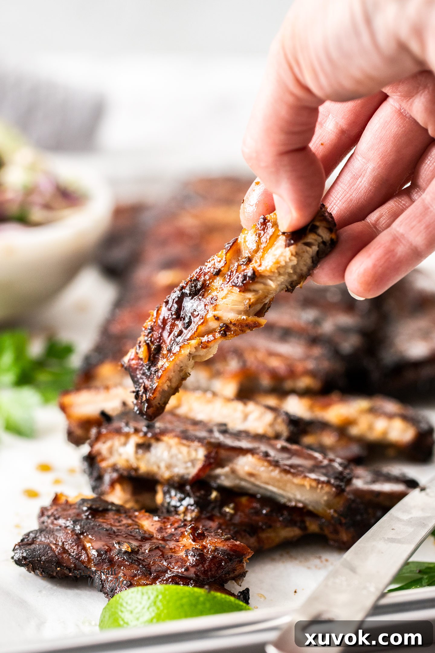 A hand holds a perfectly cooked, glazed honey garlic pork rib, showcasing its tenderness and sticky coating. The meat is visibly soft, promising to fall off the bone.