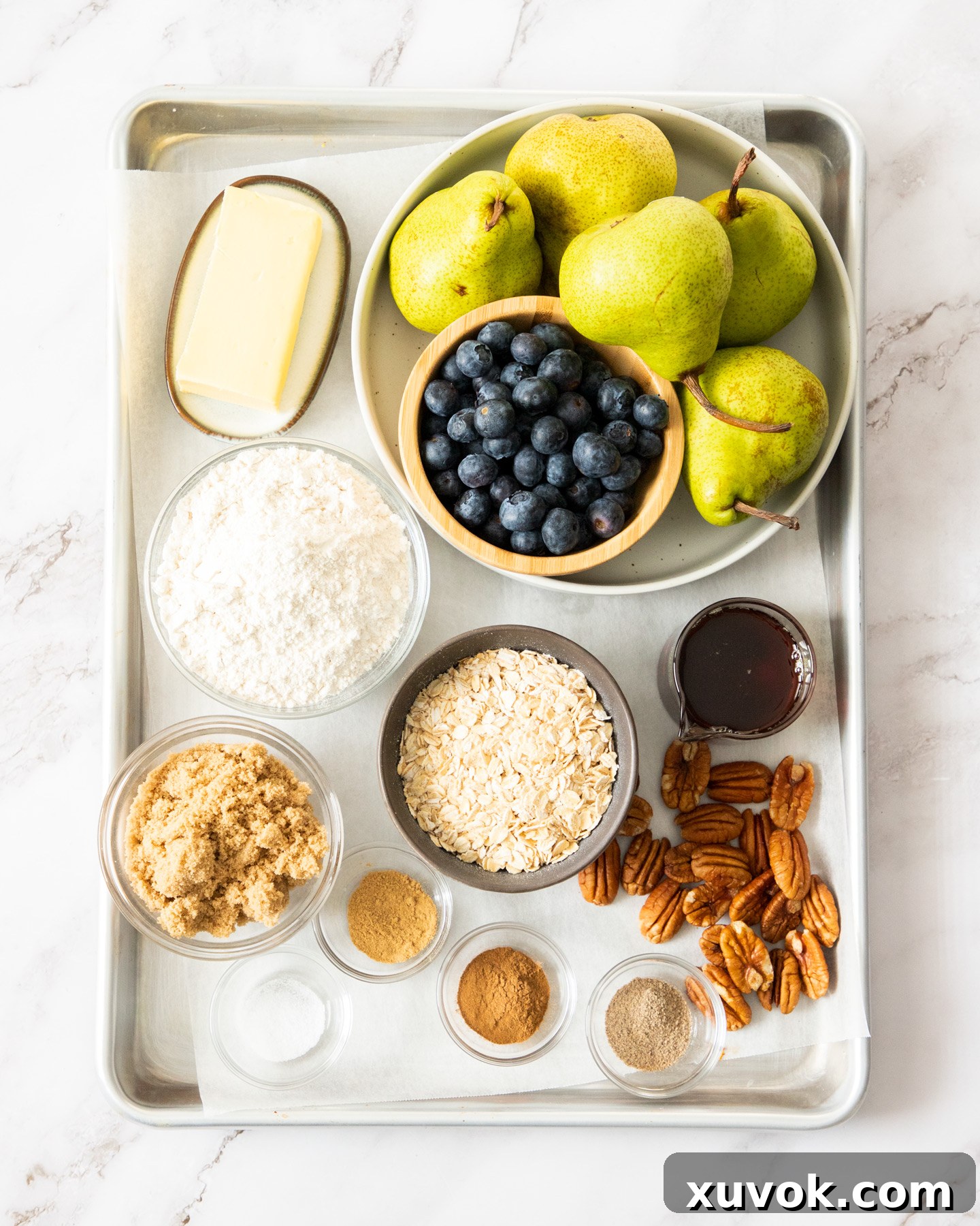 Fresh ingredients for pear and blueberry crumble laid out on a baking tray, including pears, blueberries, oats, flour, and spices.