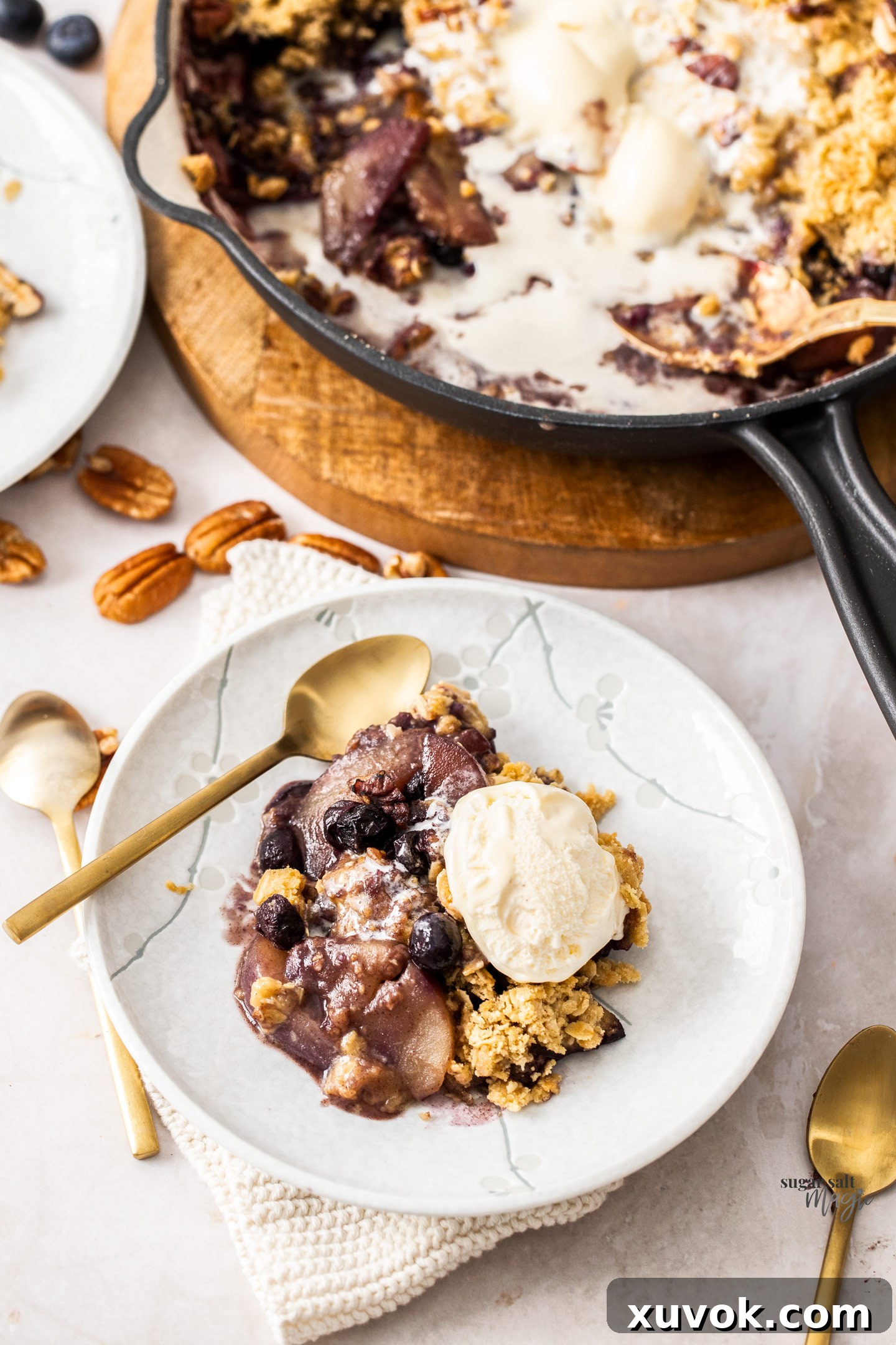A slice of warm pear and blueberry crumble on a white plate, served with a melting scoop of vanilla ice cream.
