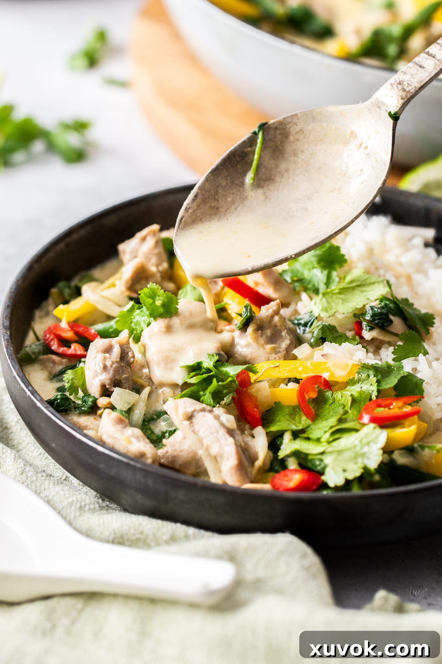 A spoon drizzling sauce over a bowl of green chicken curry. A close-up shot of a spoon drizzling rich, creamy Thai Green Chicken Curry sauce over a serving in a bowl, highlighting the luscious texture and vibrant color of the dish. The steam rising from the bowl suggests it's freshly prepared and piping hot.