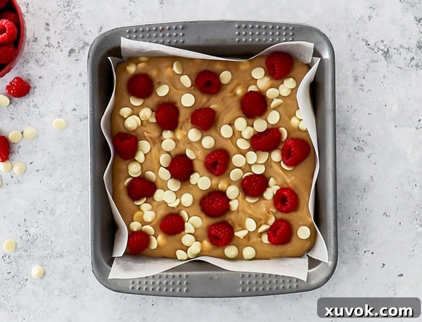 Blondie batter in a square baking pan topped with an abundance of white chocolate chips and fresh raspberries, ready for baking.