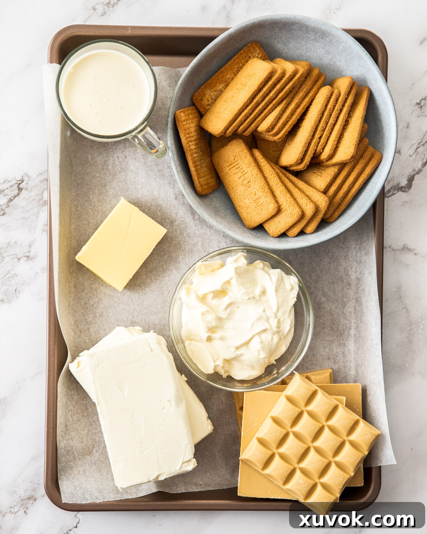 A neat arrangement of Caramilk cheesecake ingredients, including biscuits, butter, cream cheese, sour cream, Caramilk chocolate, and heavy cream on a baking tray.