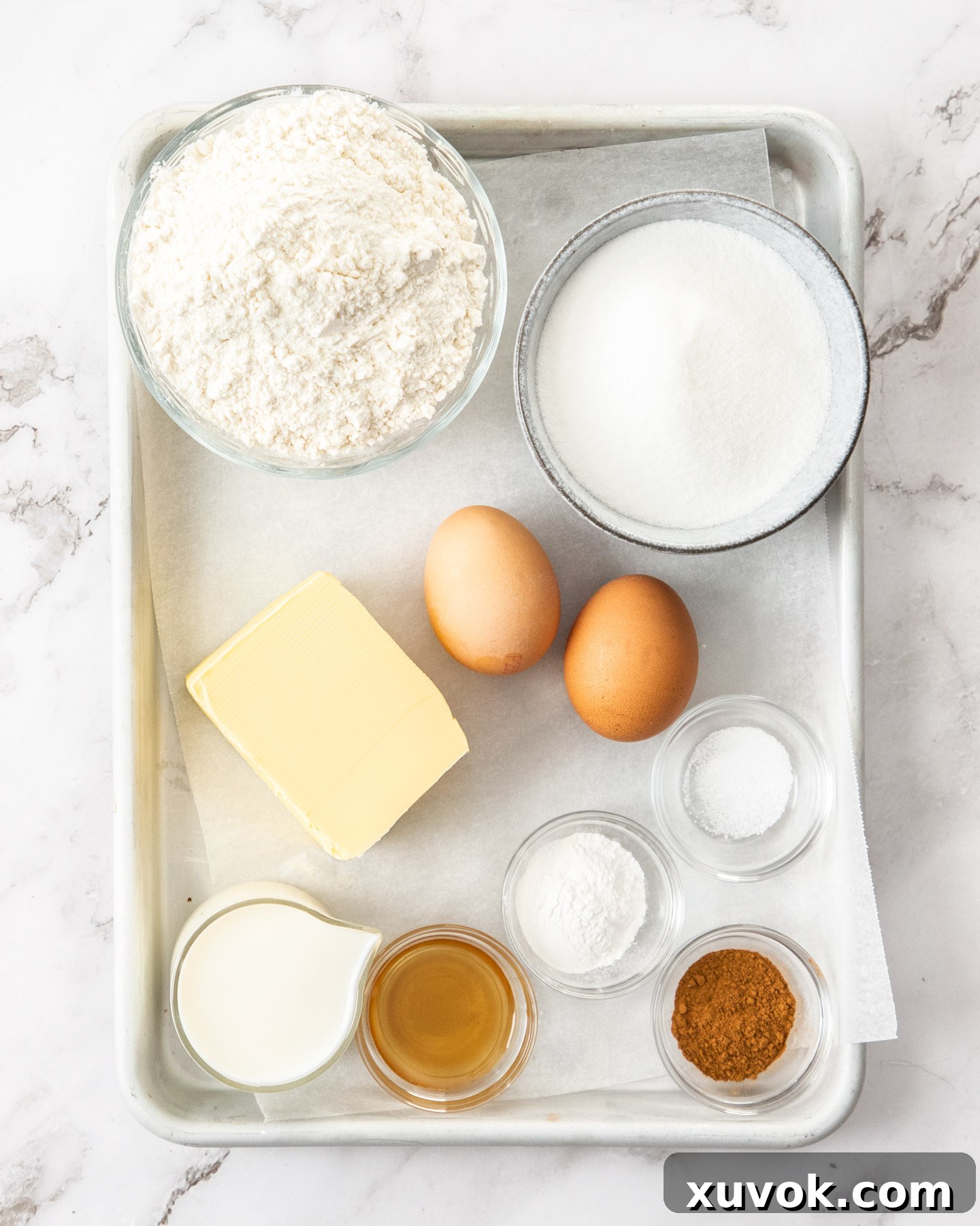 A beautifully arranged flat lay of all the fresh ingredients required to make a cinnamon teacake, including flour, butter, sugar, eggs, milk, and cinnamon sticks, laid out on a rustic baking tray.