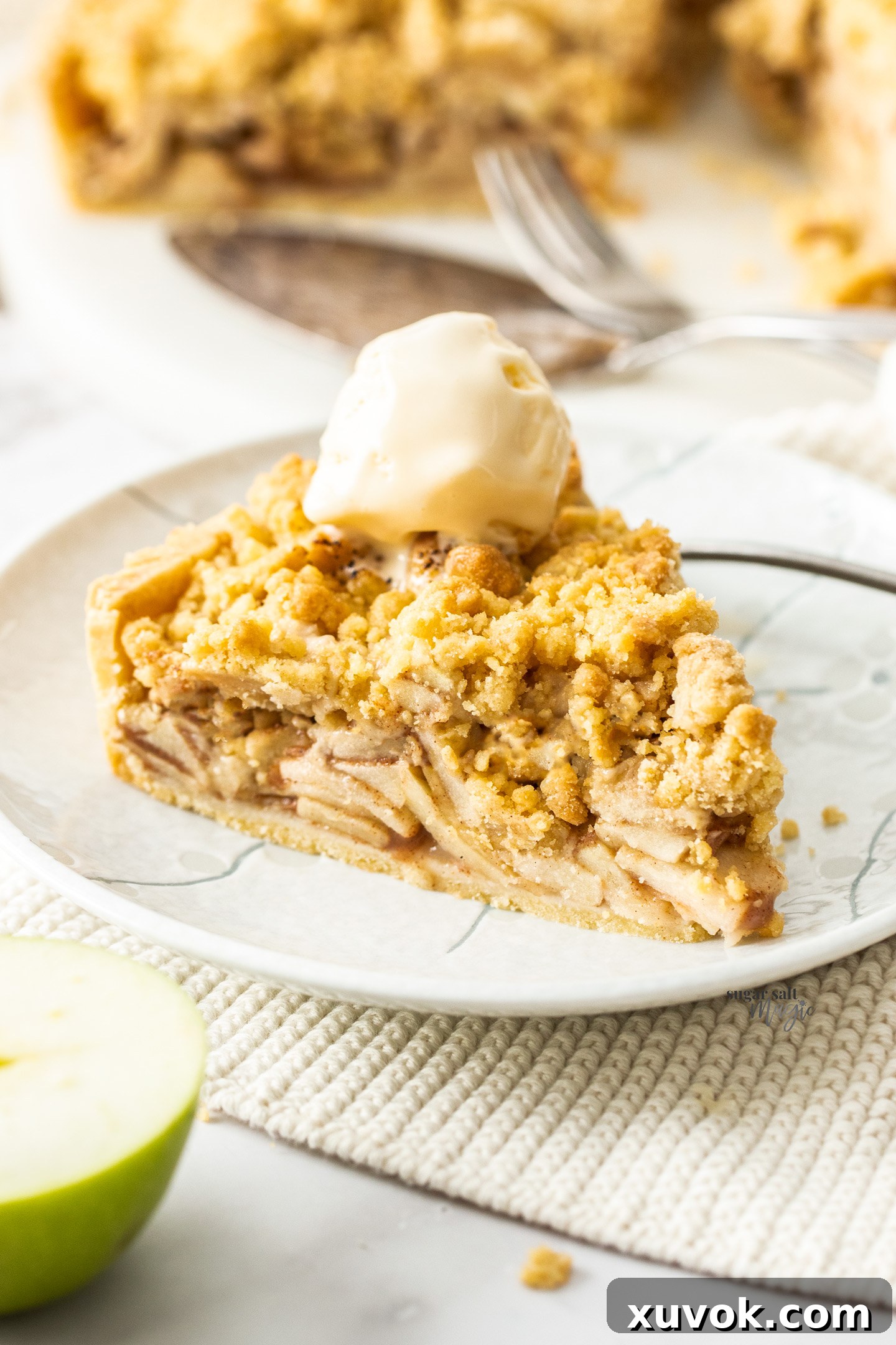 A generous slice of golden brown apple crumble tart, topped with a dollop of vanilla ice cream, sits on a rustic wooden board. The tart features a flaky crust, tender apple filling, and a crunchy streusel topping, inviting a delightful dessert experience.
