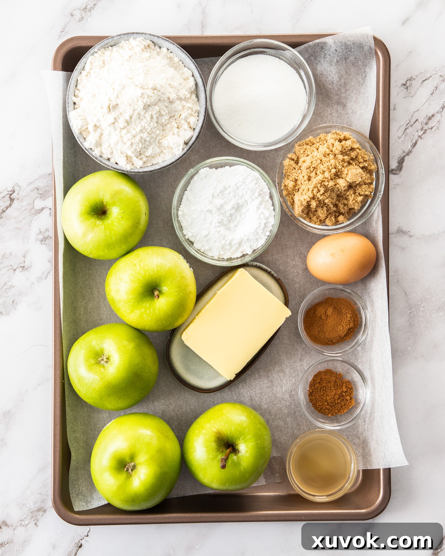 A collection of fresh Granny Smith apples, sticks of butter, bags of flour, and various sugars and spices arranged neatly on a baking tray, ready for making an apple crumble tart.