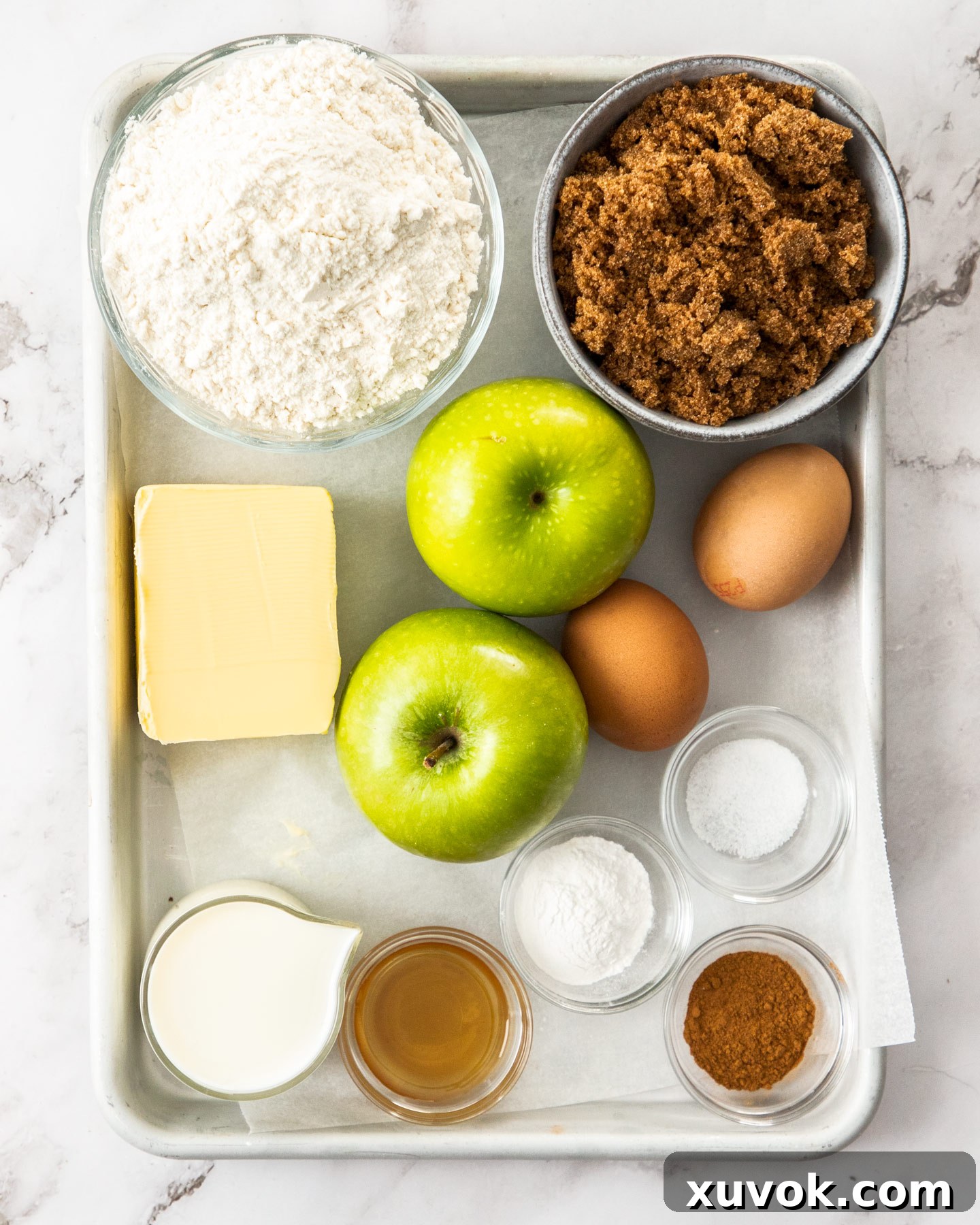 Various ingredients for an apple tray bake laid out neatly on a wooden baking tray, including fresh apples, flour, butter, sugar, and spices.