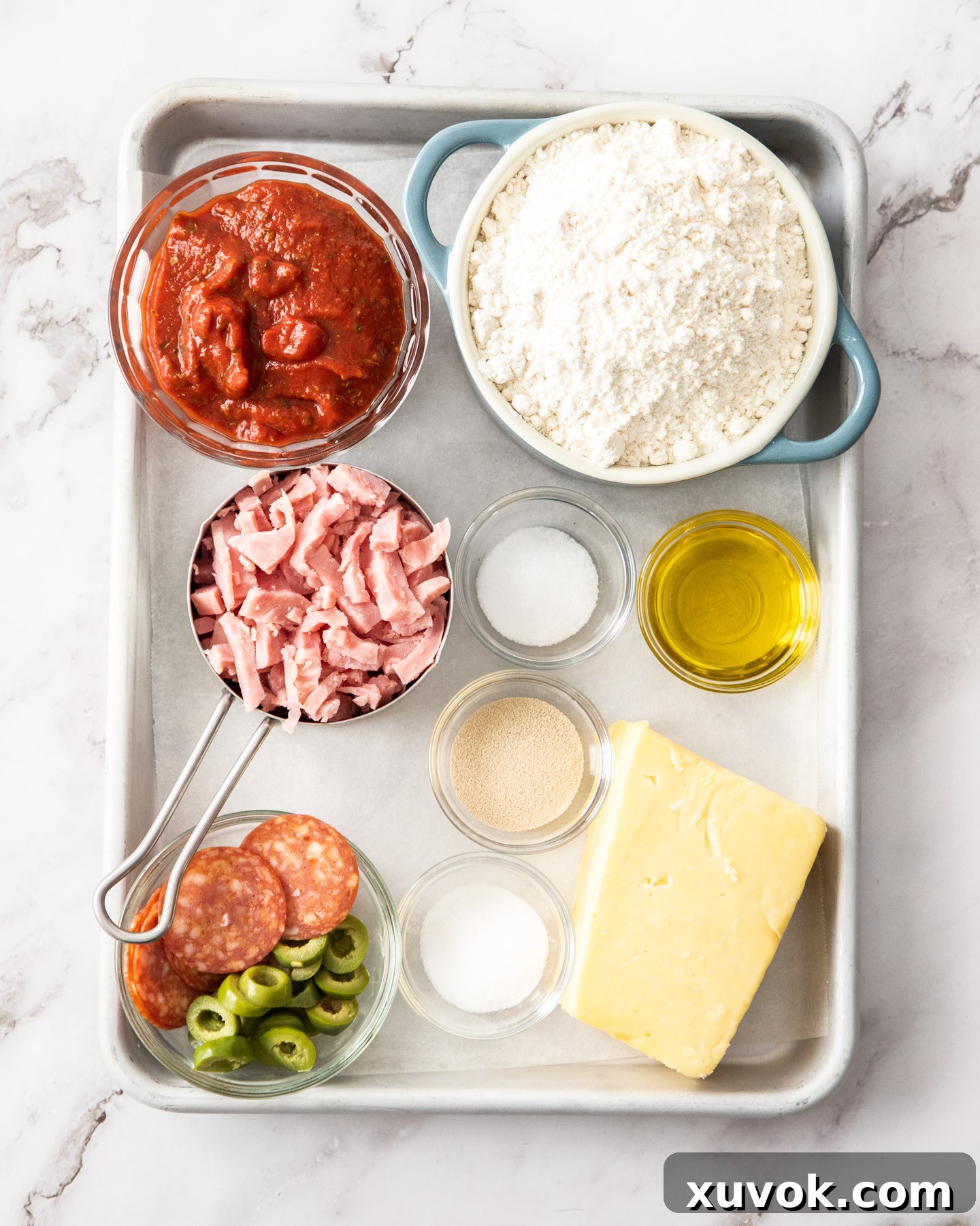 A selection of fresh ingredients laid out on a baking tray, including flour, yeast, cheese, ham, and a bowl of pizza sauce, ready for making pizza scrolls.