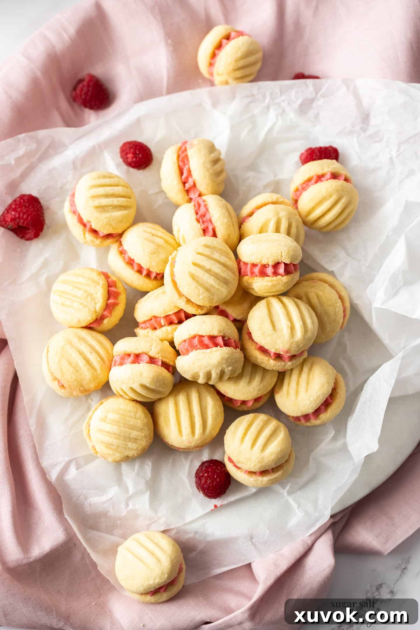 A batch of delicate Raspberry Melting Moments cookies, filled with luscious pink raspberry buttercream, artfully arranged on a sheet of baking paper from a top-down perspective.