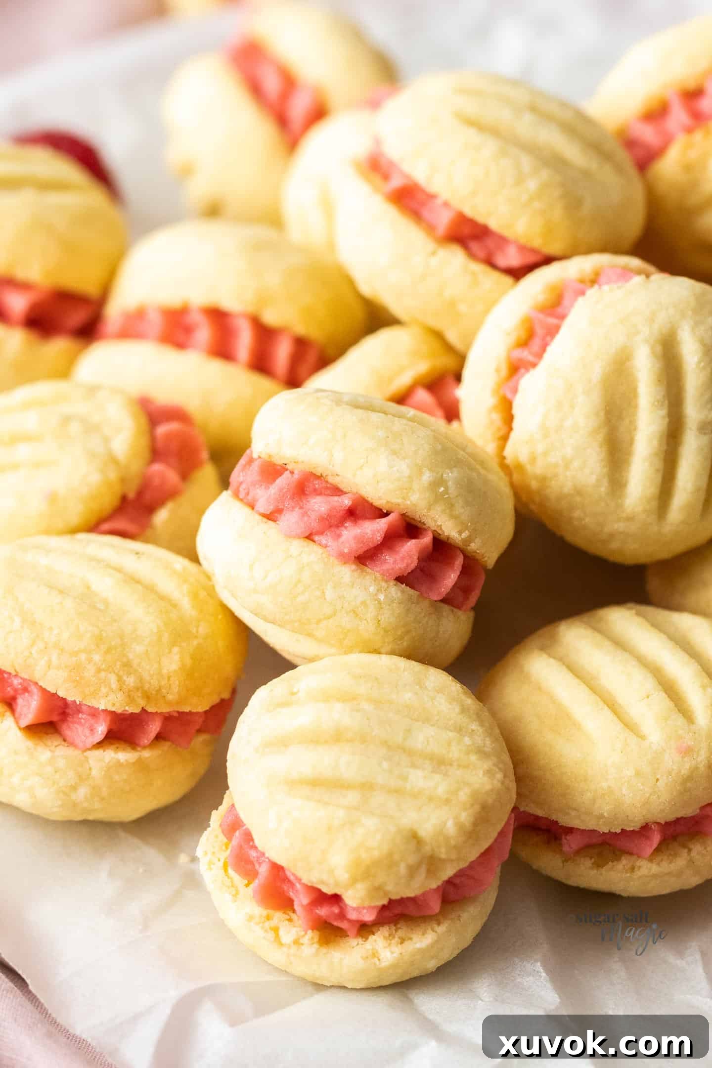 A close-up view of a small stack of Raspberry Melting Moments cookies, showcasing their delicate shortbread texture and vibrant raspberry buttercream filling, resting on baking paper.