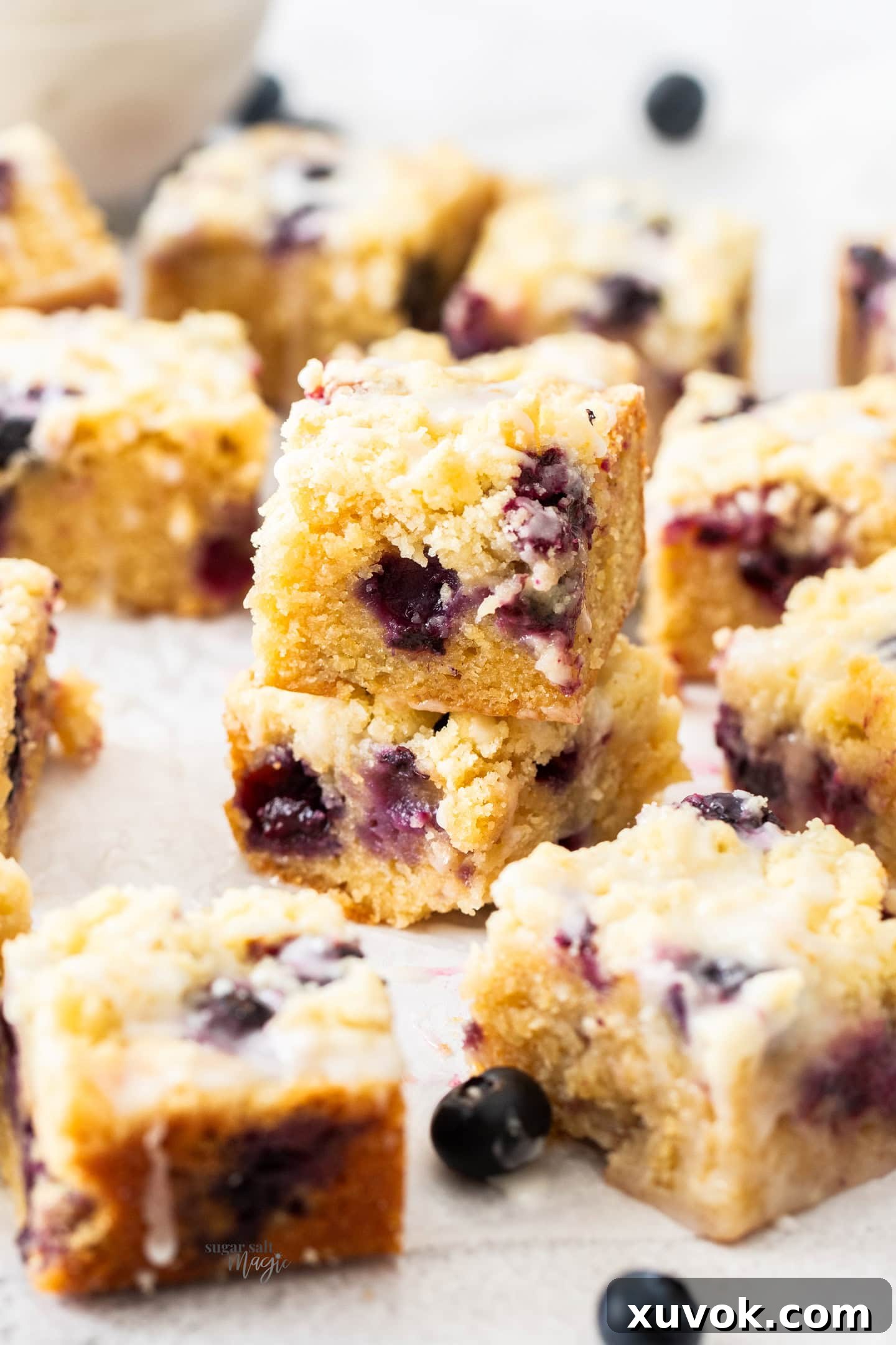 Two chewy blueberry blondies with lemon glaze, stacked, with more surrounding them on a white surface.
