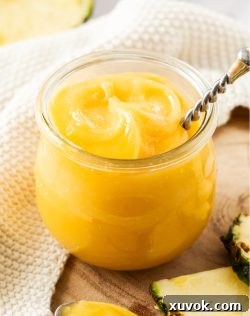 A glass jar filled with luscious pineapple curd, with fresh pineapple slices in the foreground.
