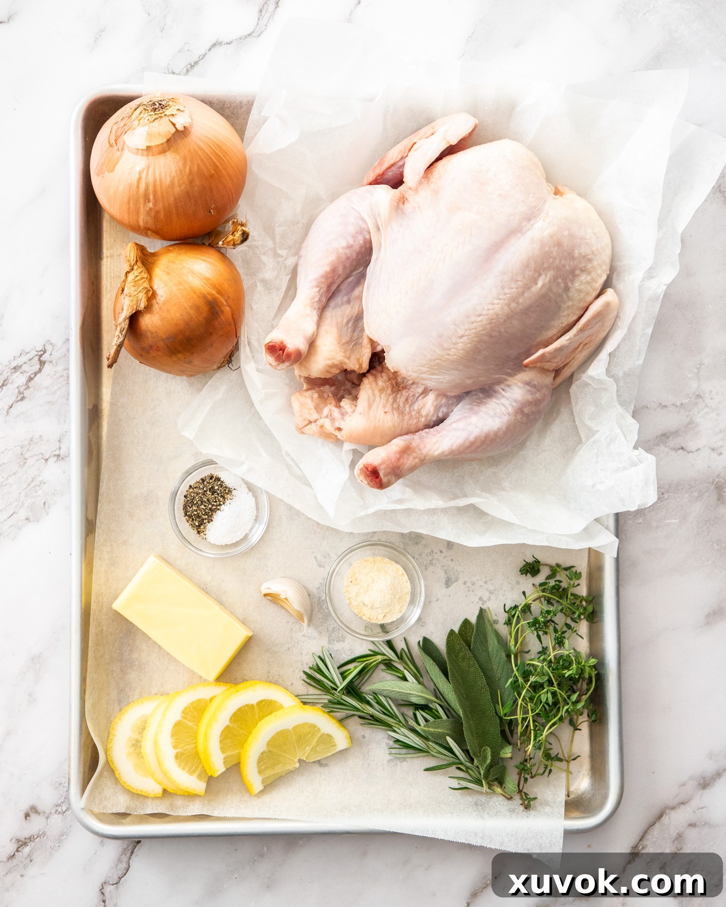 Ingredients for herb roasted chicken arranged on a baking tray, including a whole chicken, fresh herbs, lemon, garlic, and onions.