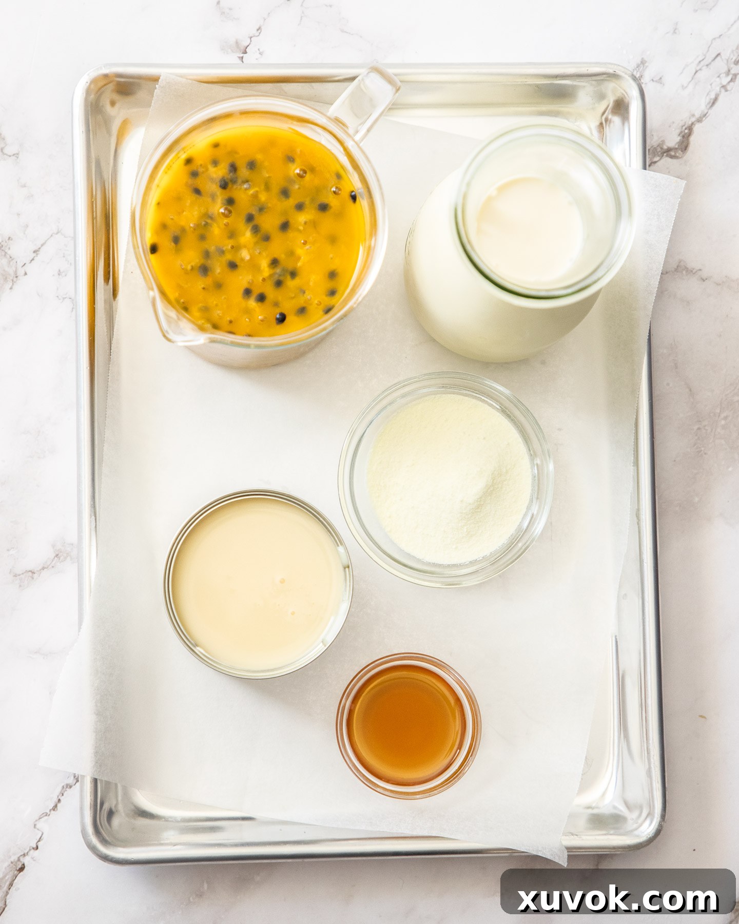 A collection of ingredients for passion fruit ice cream displayed on a wooden tray, including fresh passion fruit, a tin of condensed milk, cream, vanilla extract, and milk powder.