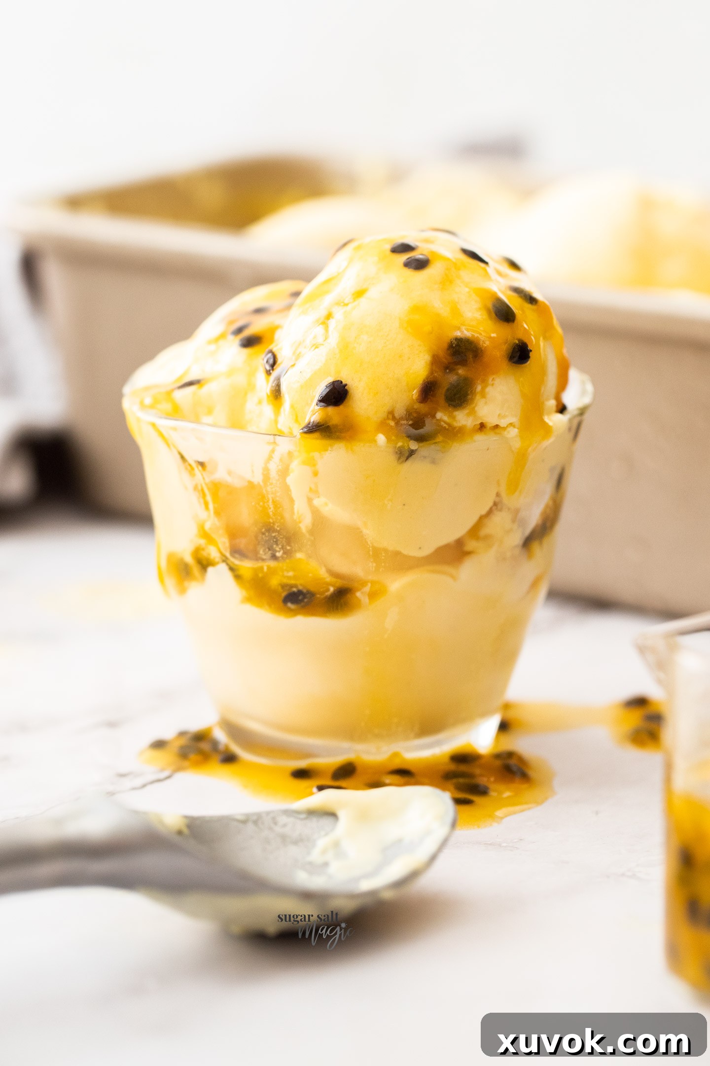 Close-up of three scoops of creamy passion fruit ice cream served in a clear glass, garnished with fresh passion fruit seeds and a mint leaf.