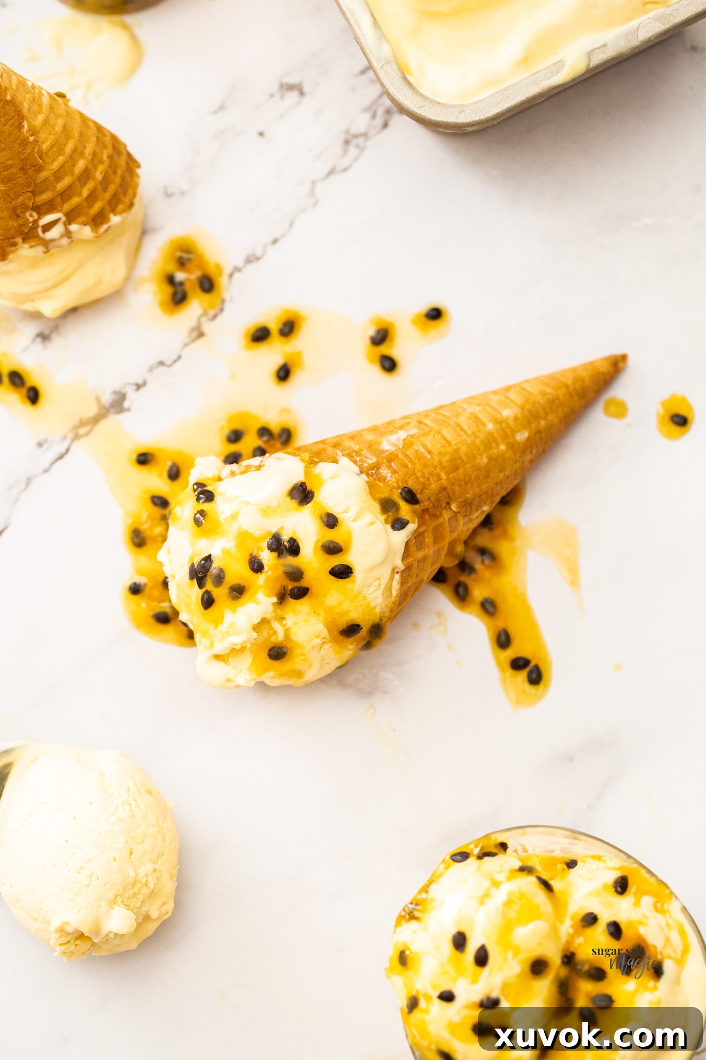 A crisp waffle cone overflowing with generous scoops of golden passion fruit ice cream, with a blurred background showing more ice cream in a freezer tub.