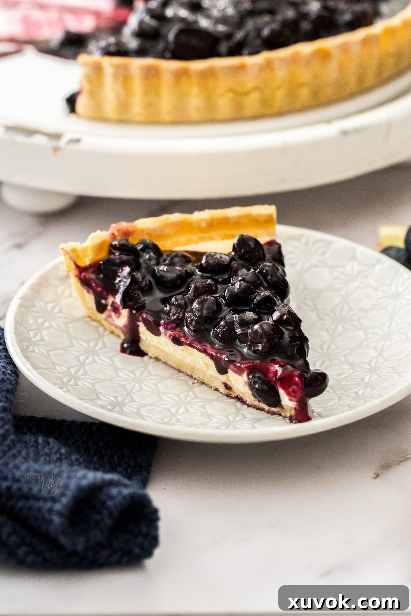 A beautifully presented slice of blueberry cheese tart on a white dessert plate, showing the distinct layers of crisp crust, creamy cheesecake filling, and glossy blueberry compote topping.