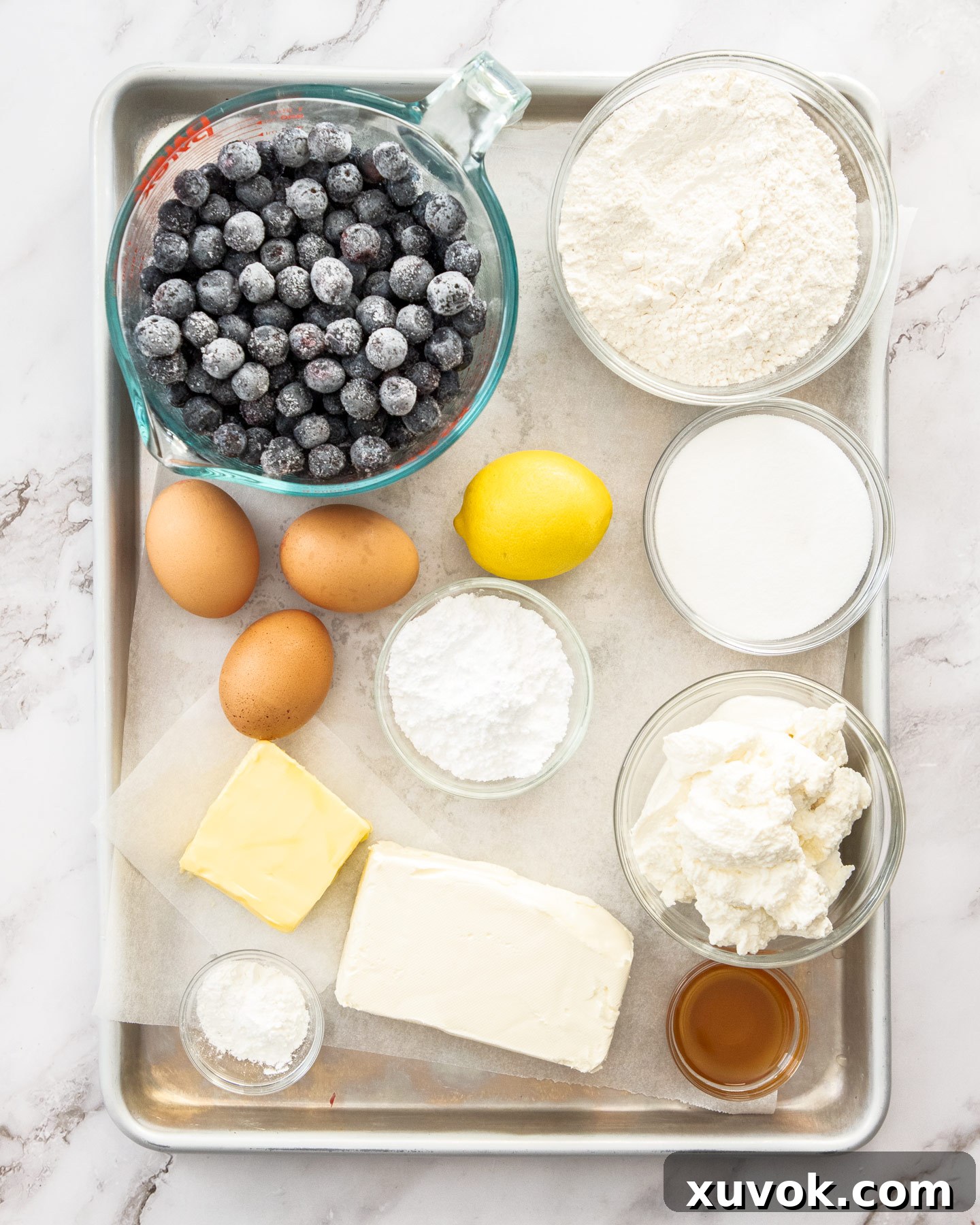 A collection of fresh ingredients for blueberry cheese tart, including butter, flour, eggs, cream cheese, ricotta, lemons, and blueberries, neatly arranged on a baking tray.