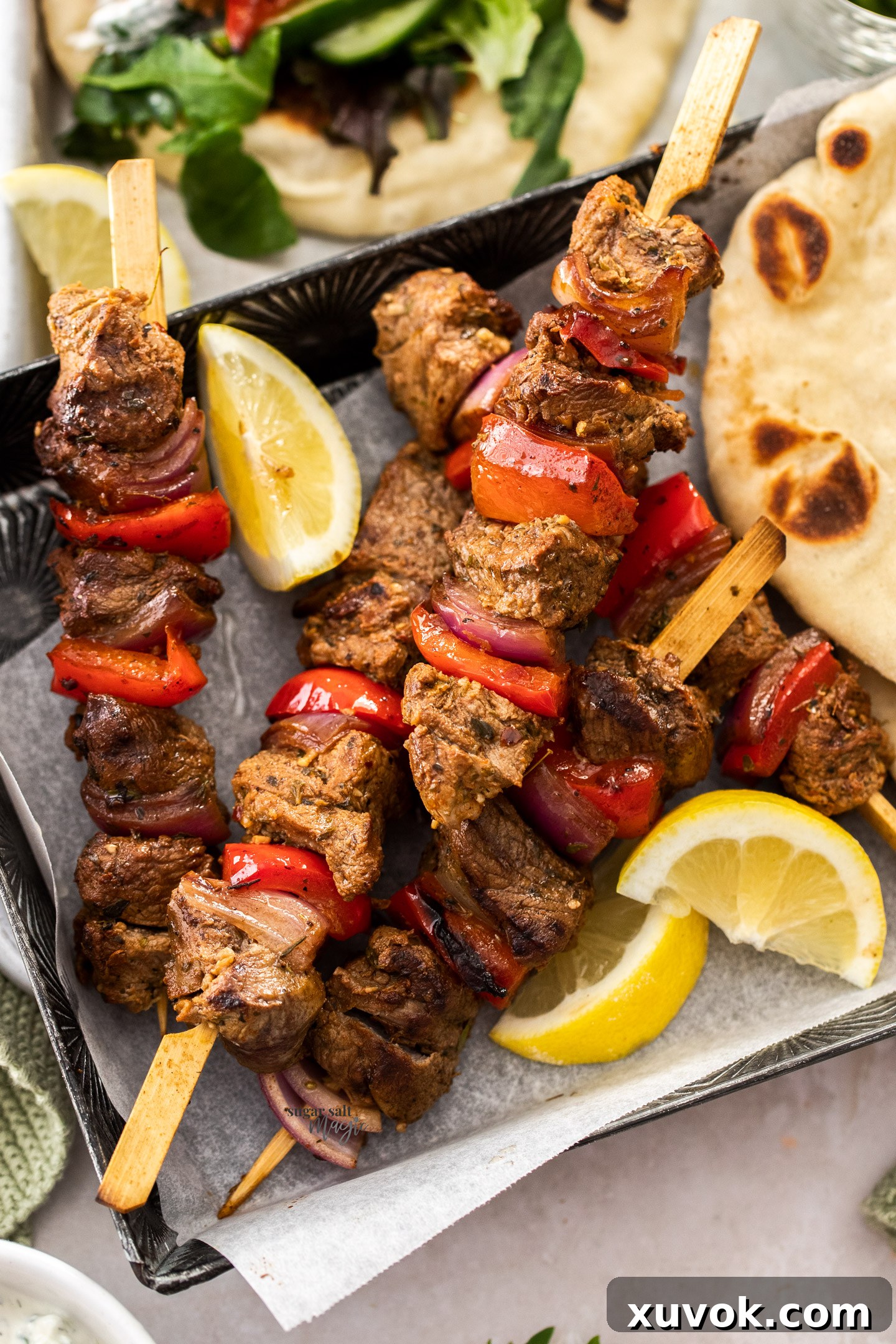 Five perfectly grilled Greek lamb kebabs, skewered with onions and bell peppers, are laid out on a dark serving tray, garnished with fresh herbs.