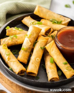 A batch of crispy homemade Chinese spring rolls arranged on a plate with a side of dipping sauce.