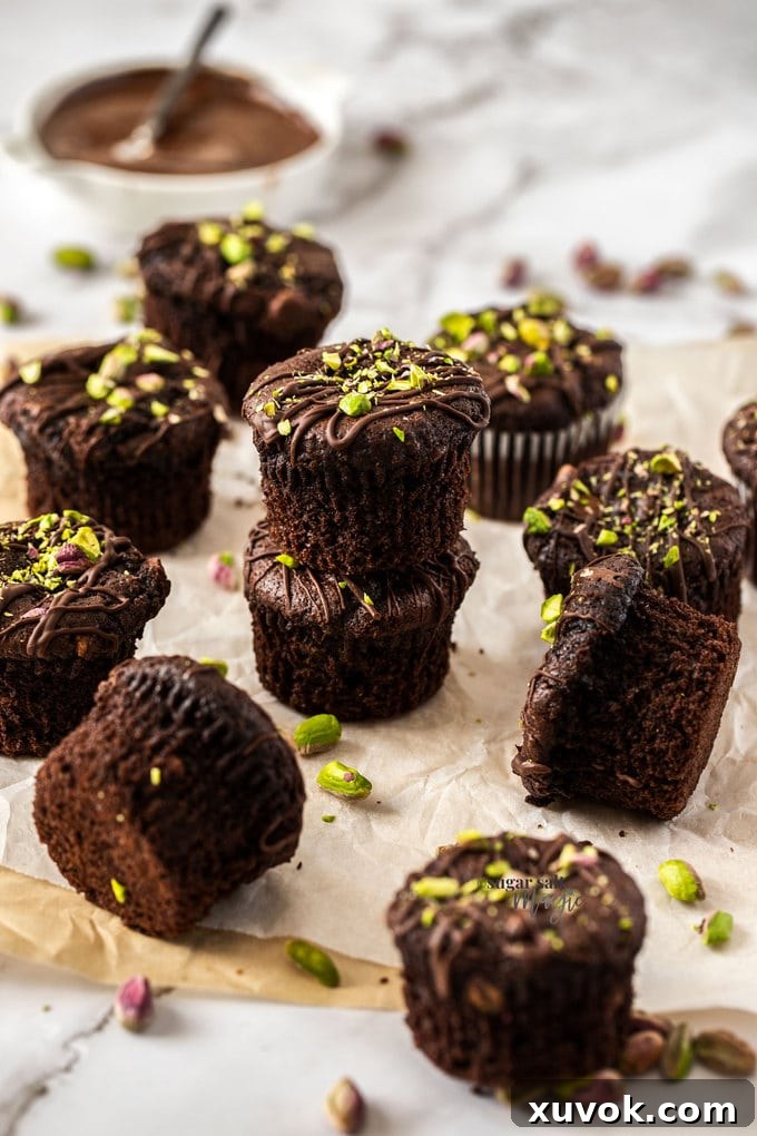 A glorious batch of freshly baked Pistachio Chocolate Chip Muffins cooling on a sheet of baking paper, generously scattered with bright green pistachios. Each muffin boasts a perfectly domed top and hints of melted chocolate.