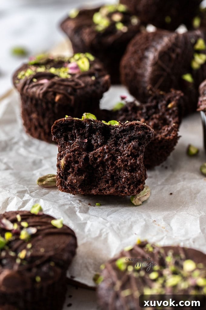 A close-up shot of a warm, rich chocolate muffin on a sheet of baking paper, its top glistening with melted chocolate and adorned with bright green pistachio pieces. More muffins are blurred in the background, creating an inviting scene.