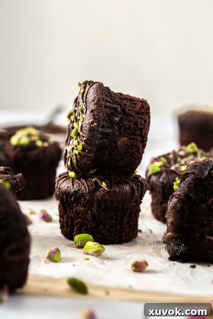 Two exquisitely stacked chocolate pistachio muffins, richly drizzled with melted chocolate and adorned with crushed pistachios. More muffins and loose pistachios are spread around, inviting you to indulge in this delightful treat.