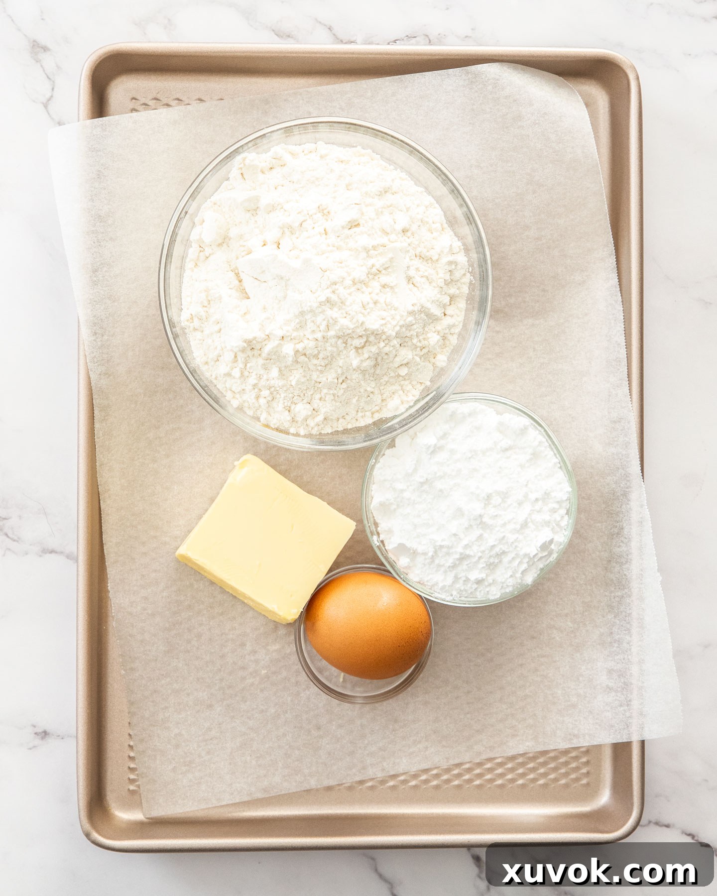 A flat lay photograph displaying the four key ingredients for tartlet shells: flour, icing sugar, cubed butter, and a cold egg, arranged neatly on a baking tray.