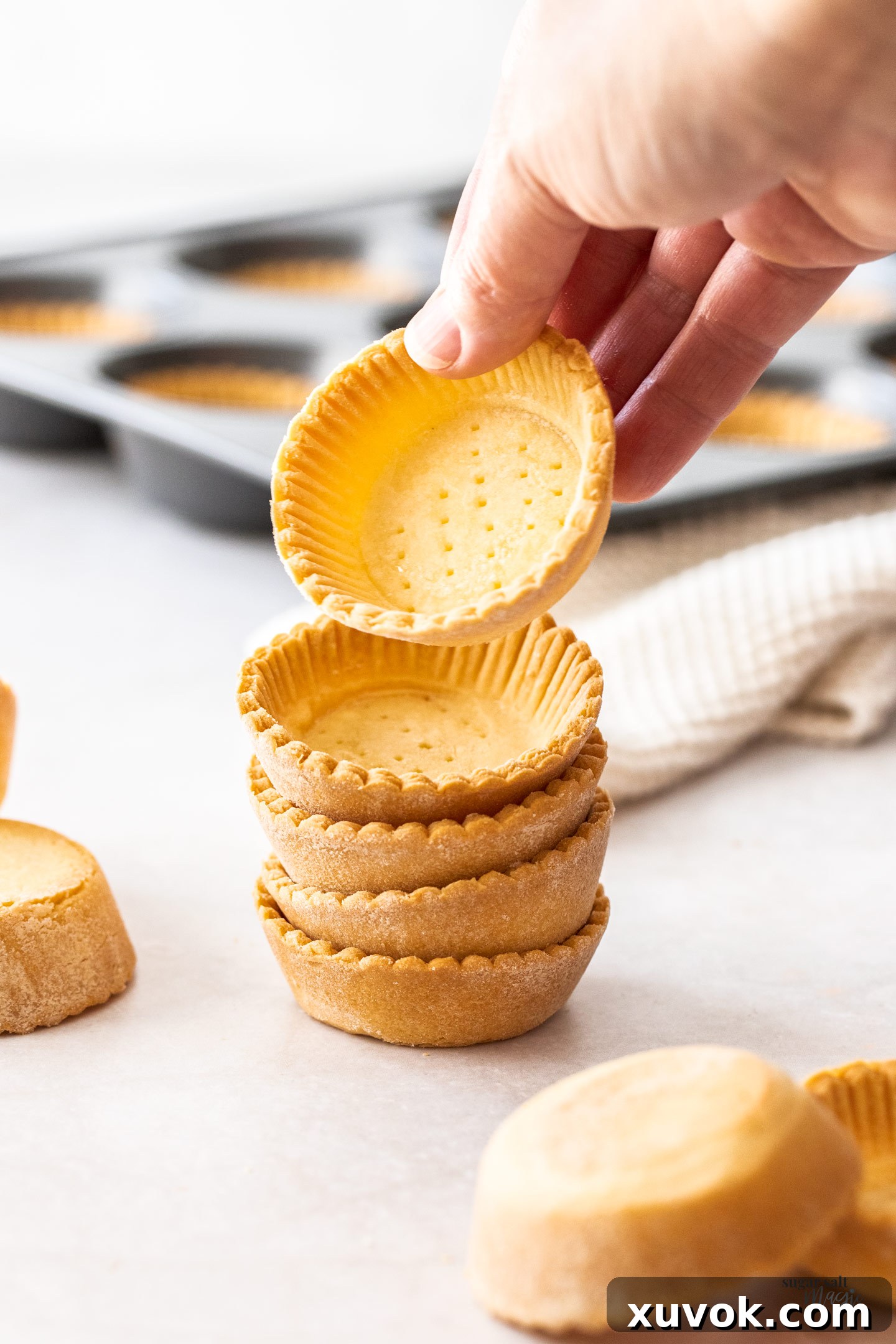 A hand reaching for a tartlet shell from a neatly stacked pile of five, highlighting their perfect form and texture.